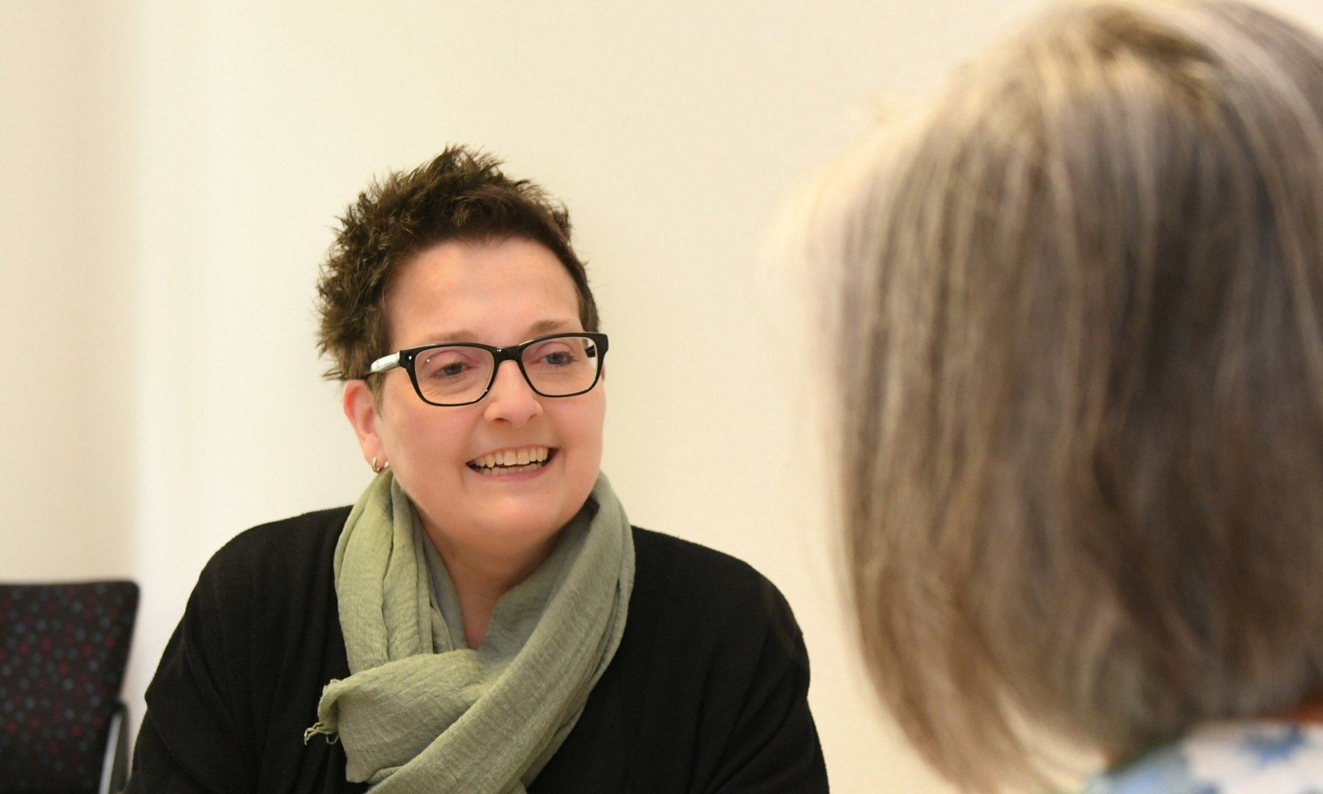 Frau Sitz an einem Schreibtisch mit einem Blatt in der Hand und berät eine andere Frau, die mit dem Rücken zur Kamera sitzt