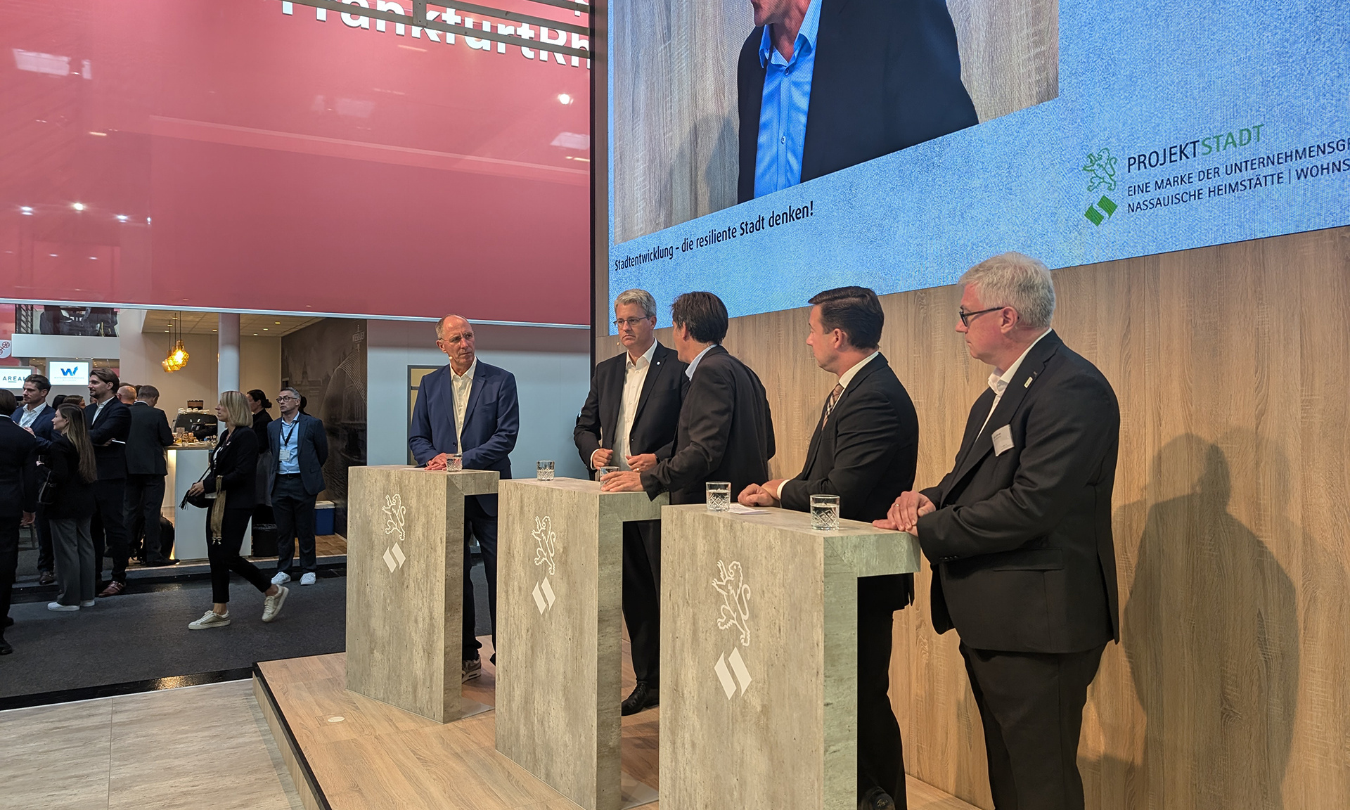 Fünf Männer hinter Rednerpulten im Gespräch miteinander, einer von ihnen wird oberhalb auf einem Screen gezeigt.