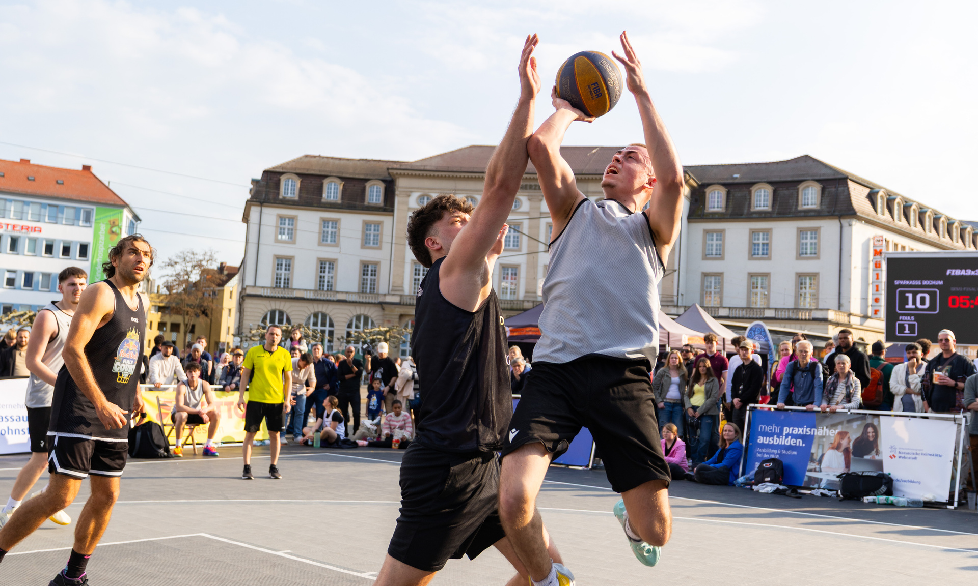 Zwei junge Männer im Zweikampf mit dem Ball unter dem Basketballkorb. Einer befindet sich im Sprung für einen Korbleger.