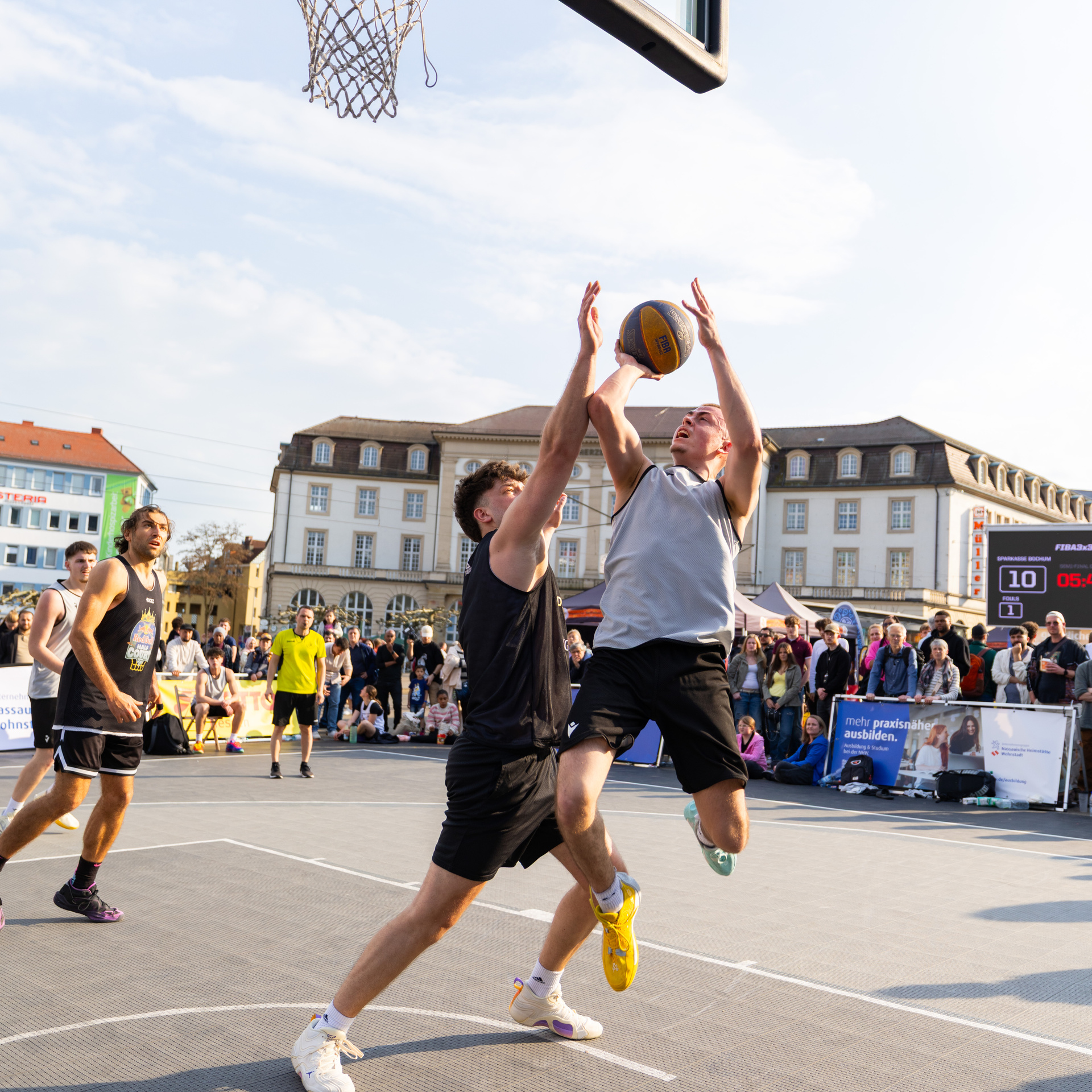 Zwei junge Männer im Zweikampf mit dem Ball unter dem Basketballkorb. Einer befindet sich im Sprung für einen Korbleger.