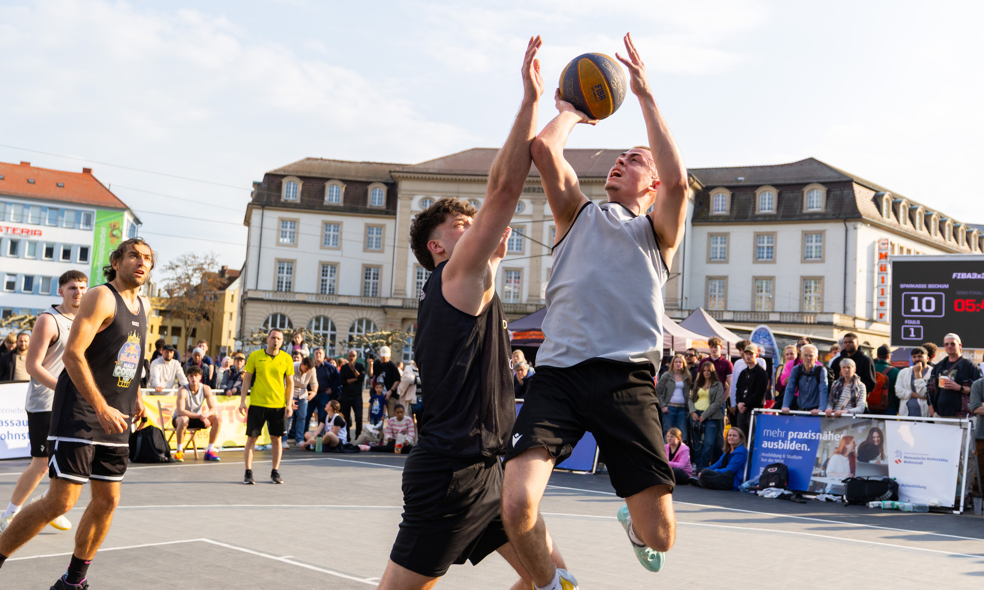 Zwei junge Männer im Zweikampf mit dem Ball unter dem Basketballkorb. Einer befindet sich im Sprung für einen Korbleger.