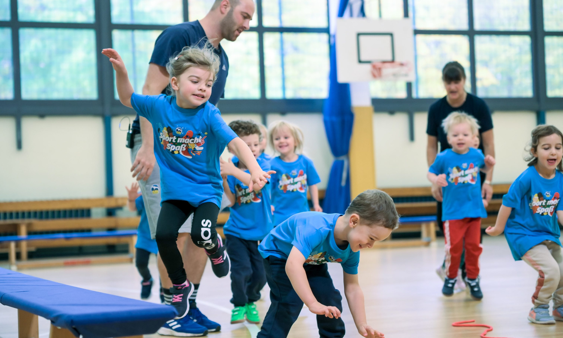 Kinder in blauen T-Shirts mit der Aufschrift "Sport macht Spaß" spielen und toben in einer Sporthalle.