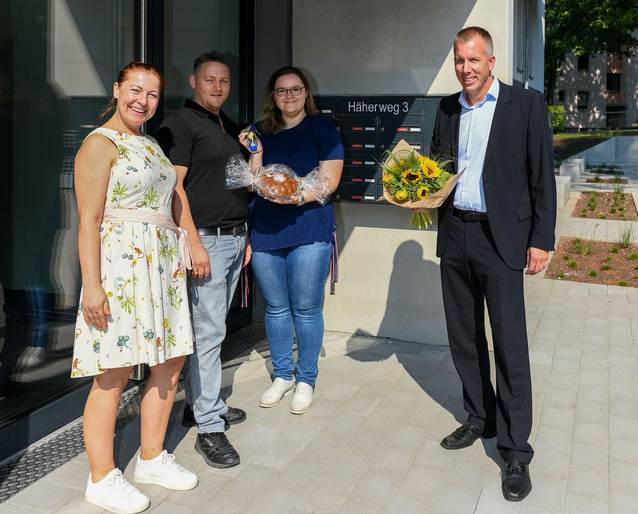 Mieterin und Mieter werden von Kundenmanagerin und Servicecenterleiter vor ihrer neuen Wohnung mit Blumenstrauß, Salz und Brot begrüßt.