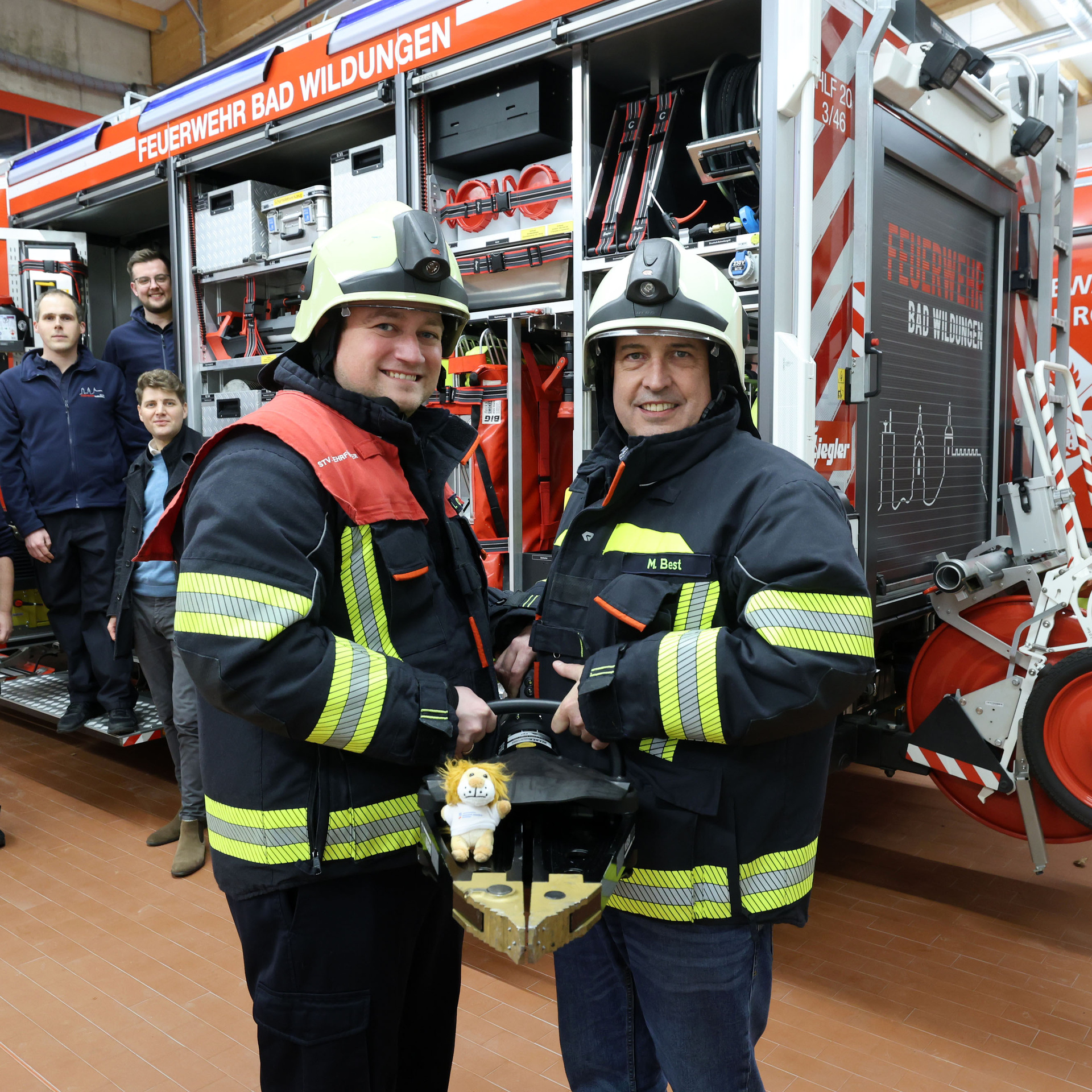 Im Vordergrund sind zwei Feuerwehrmänner mit Ausrüstung. Im Hintergrund steht ein Löschfahrzeug und weitere Personen steigen aus dem Feuerwehrauto aus.