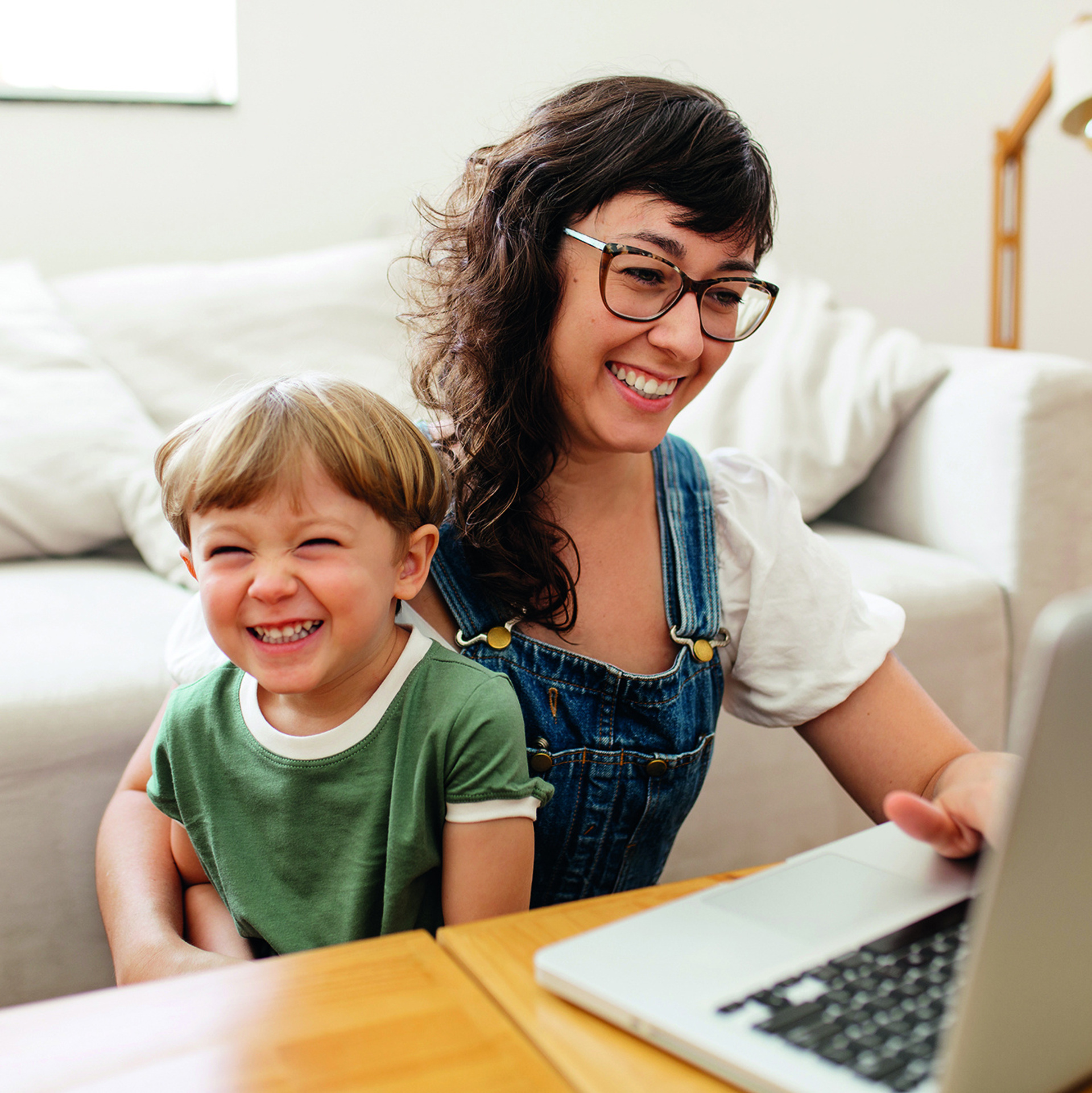 Frau mit Brille sitzt mit einem Kleinkind vor einem Laptop. Kleiner Junge lacht in die Kamera, Frau schaut in den Laptop.
