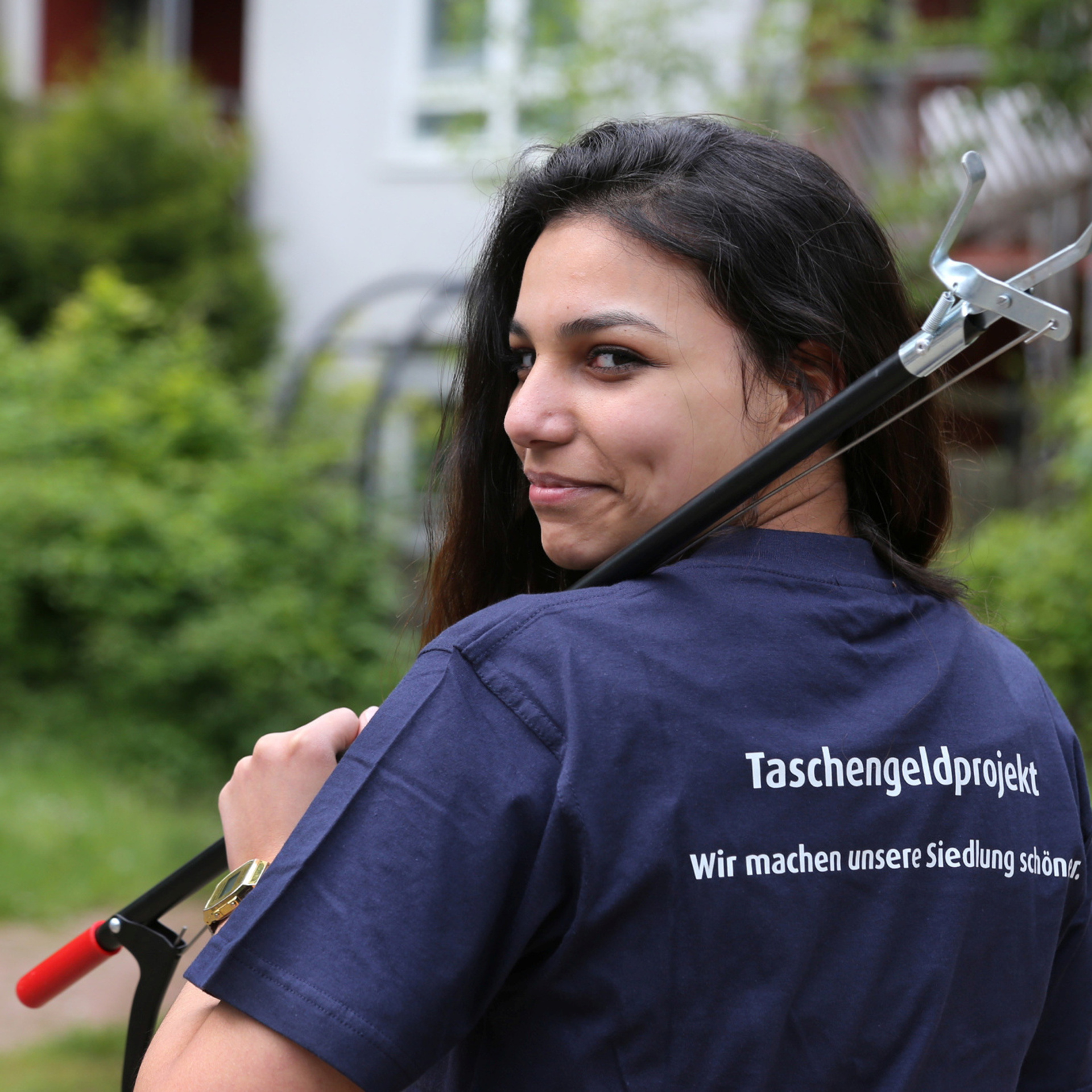 Junges Mädchen, mit einer Müllgreifzange, blickt über die Schulter. Auf dem T-Shirt auf dem Rücken steht Taschengeldprojekt.