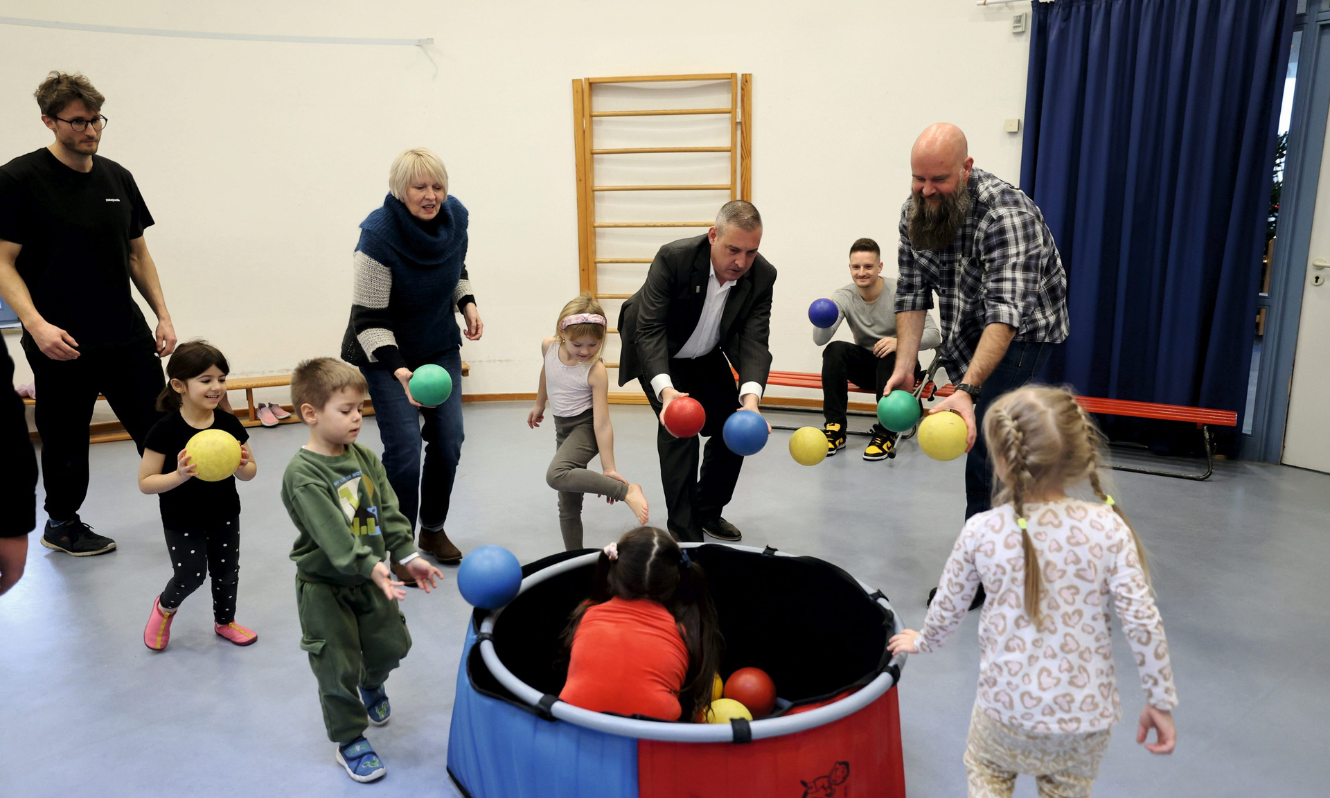 Mehrere Erwachsene spielen mit Kindern in einer Turnhalle. In der Mitte steht eine Art Bällebad, in dem auch Kinder sitzen. Alle werfen sich gegenseitig Bälle zu.