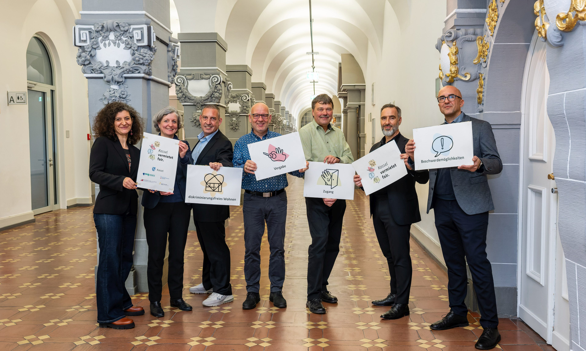Mitglieder der Initiative Kassel vermietet fair. Jeder hält eine Tafel mit einzelnen Elementen des Leitbilds in die Höhe (v.l.n.r.): Teslihan Ayalp (Leiterin Amt für Chancengleichheit), Stadtbaurätin Simone Fedderke, Sascha Holstein (NHW, Leiter Regionalcenter Kassel), Sven Hansen (Bauverein 1894), Michael Fuchs (Studierendenwerk), Uwe Gabriel (GWG), Evren Özgüvenc (Antidiskriminierungsbeauftragter).
