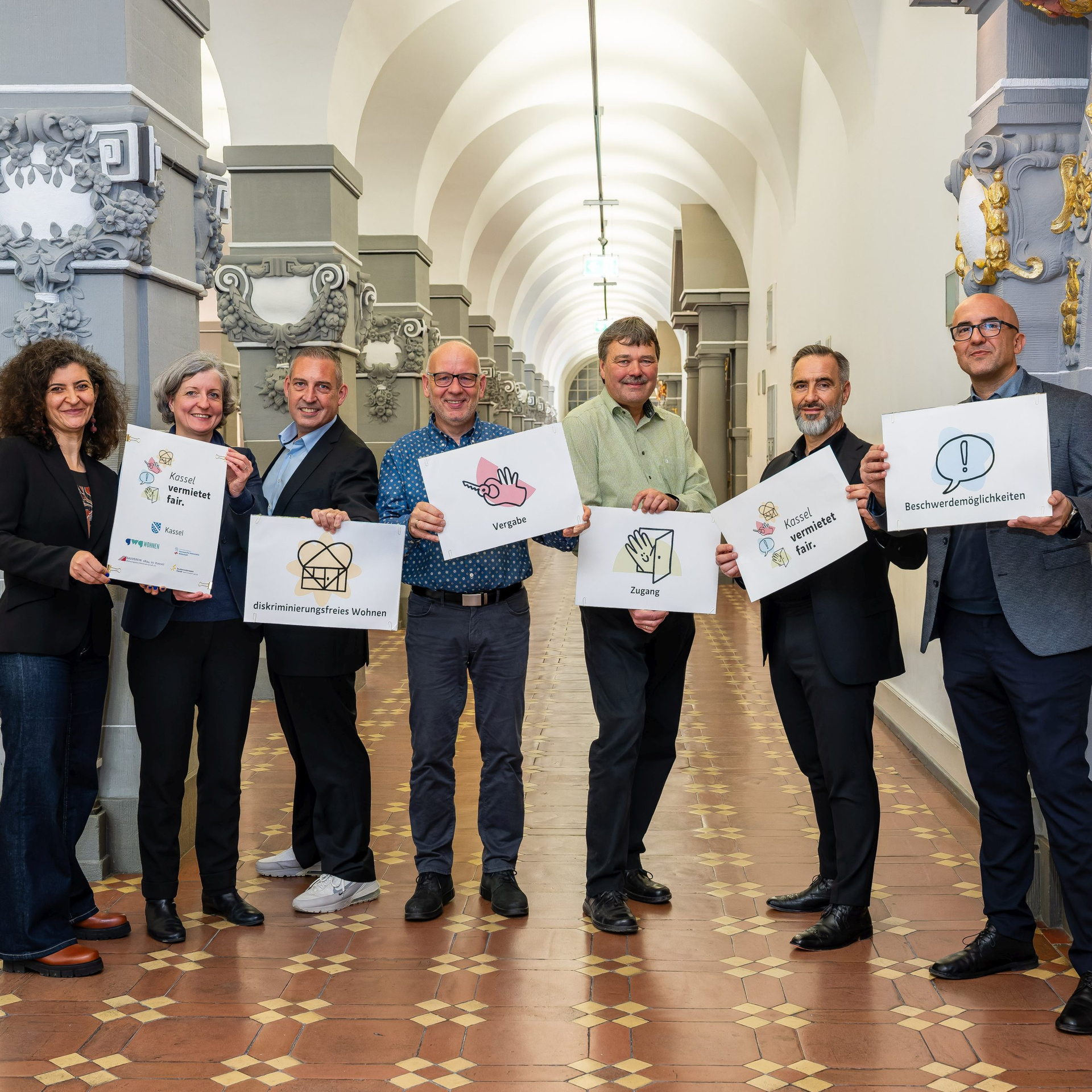 Mitglieder der Initiative Kassel vermietet fair. Jeder hält eine Tafel mit einzelnen Elementen des Leitbilds in die Höhe (v.l.n.r.): Teslihan Ayalp (Leiterin Amt für Chancengleichheit), Stadtbaurätin Simone Fedderke, Sascha Holstein (NHW, Leiter Regionalcenter Kassel), Sven Hansen (Bauverein 1894), Michael Fuchs (Studierendenwerk), Uwe Gabriel (GWG), Evren Özgüvenc (Antidiskriminierungsbeauftragter).