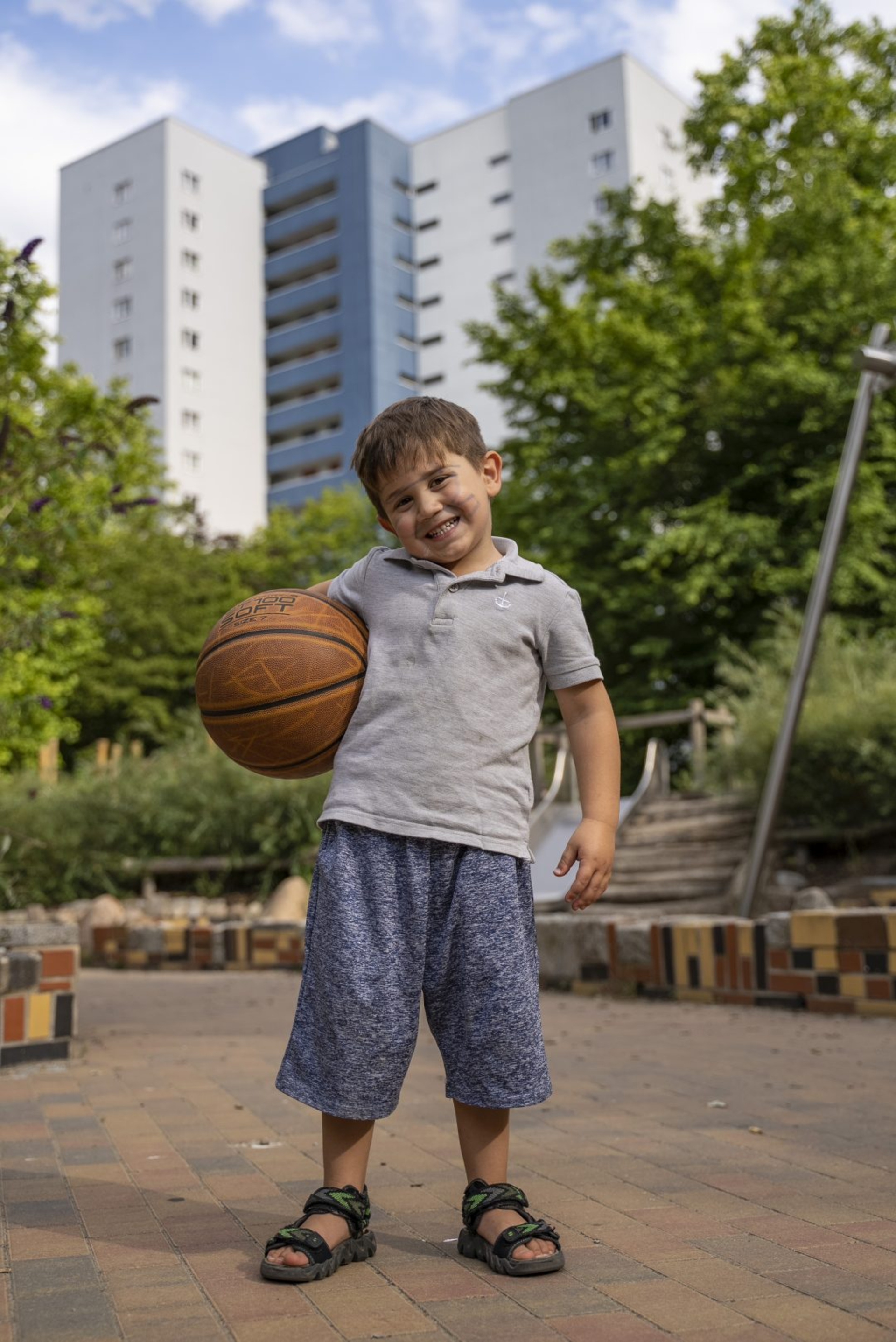 Ein kleiner Junge mit einem Basketball in der Hand steht auf einem Spielplatz vor einem Wohnhochhaus.