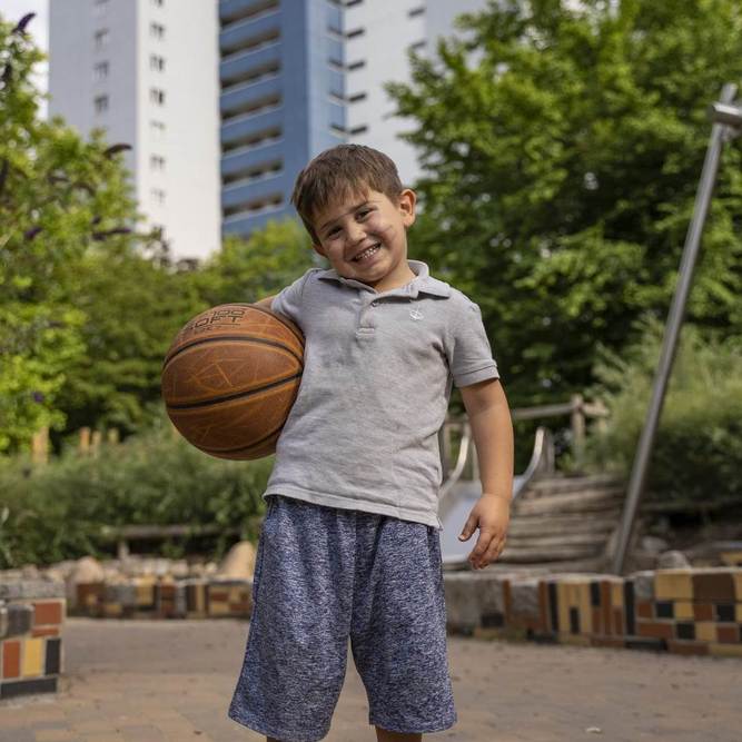 Ein kleiner Junge mit einem Basketball in der Hand steht auf einem Spielplatz vor einem Wohnhochhaus.