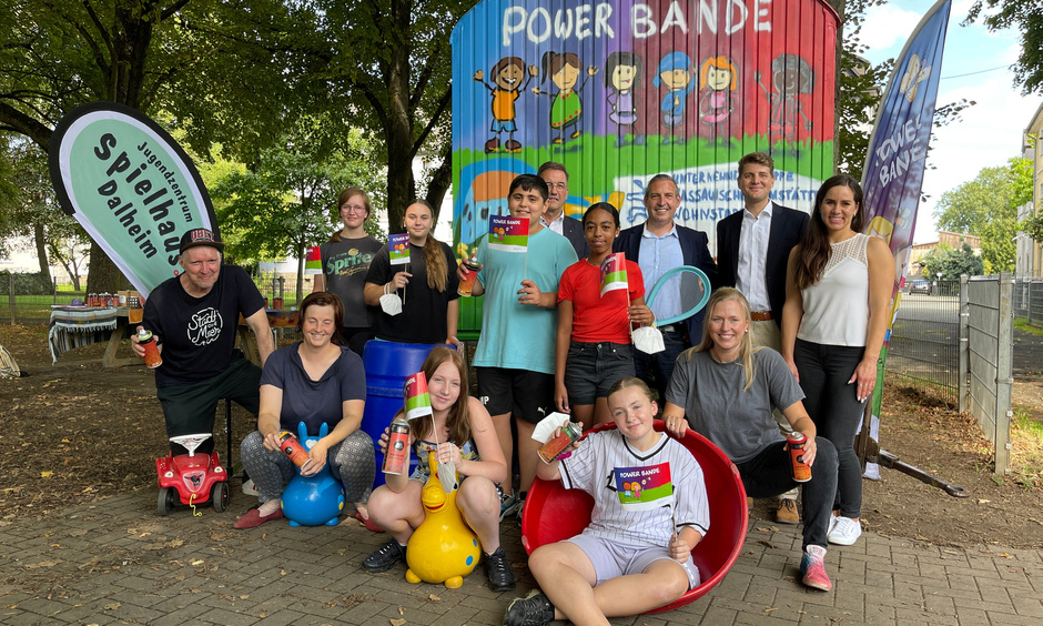 Eine Gruppe von Kindern und Erwachsenen im Freien vor einem bunten Bauwagen, alle halten Spielzeug in der Hand.