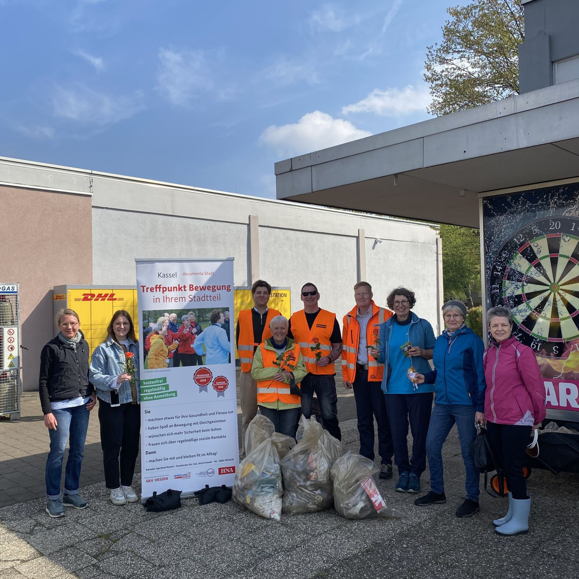Gruppe von Menschen mit gesammelten Müllbeuteln im Vordergrund. Aufsteller "Treffpunkt Bewegt"