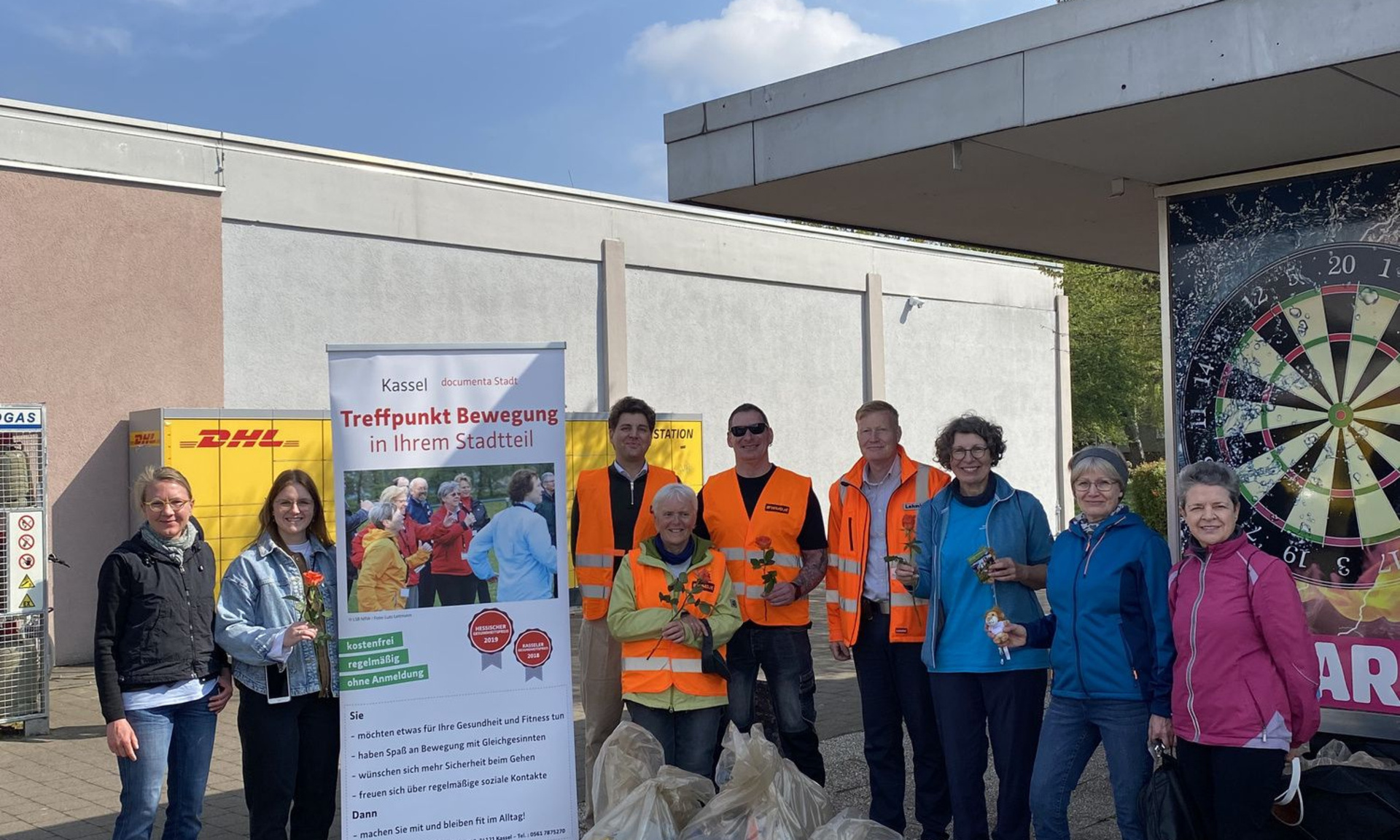 Gruppe von Menschen mit gesammelten Müllbeuteln im Vordergrund. Aufsteller "Treffpunkt Bewegt"