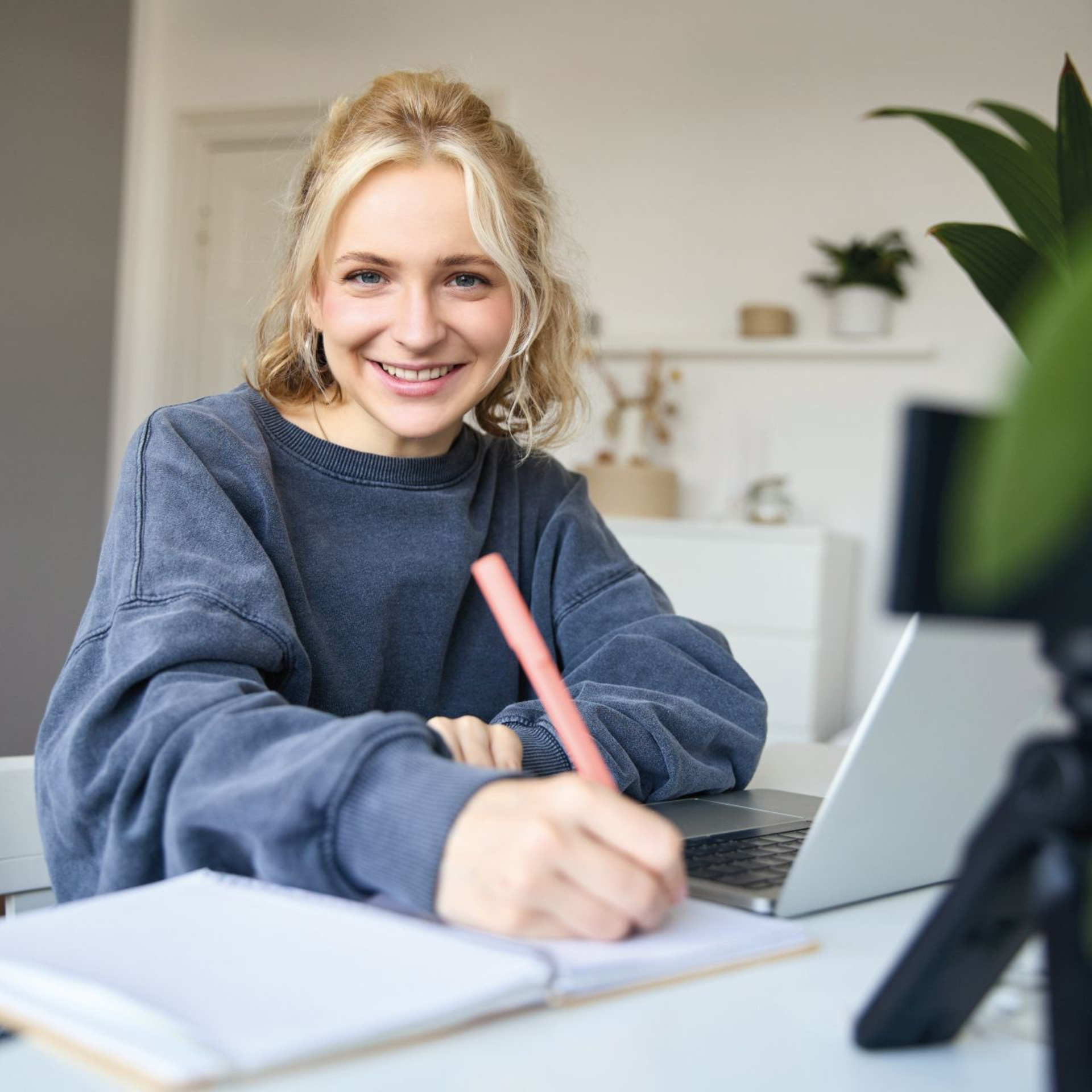 Mädchen sitzt an einem Laptop und schreibt etwas auf einen Block neben dem Laptop. Mädchen sieht in die Kamera und lacht.