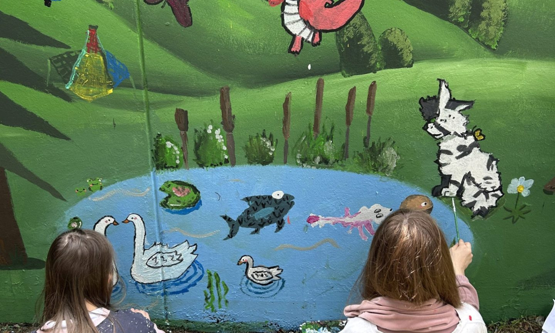 Zwei Kinder von hinten zu sehen bemalen mit bunten Wasserfarben und Pinsel eine Wand mit Tieren, Blumen, einem See und einem Regenbogen