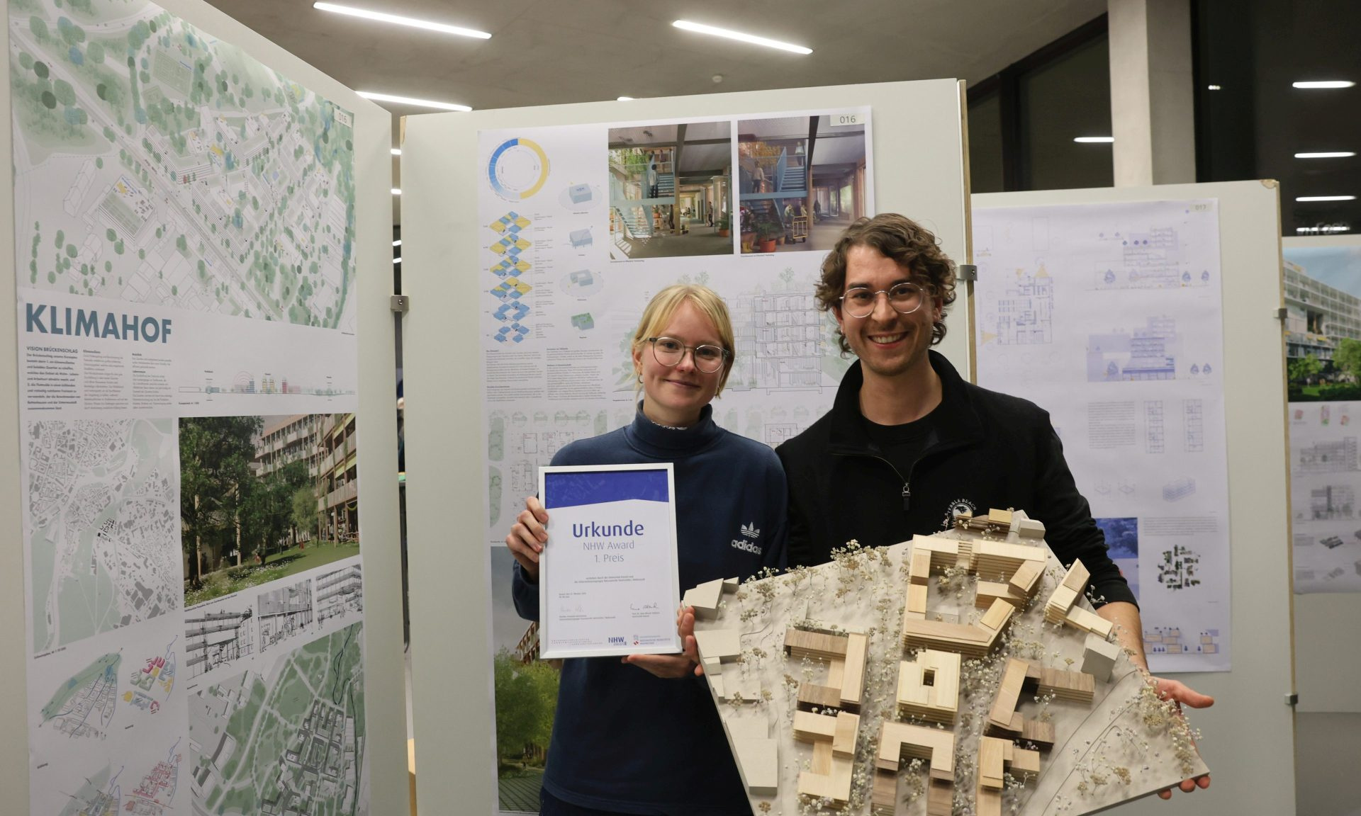Elina Klett und Tom Stehmann belegten mit ihrem Projekt Klimahof einen der beiden ersten Plätze. Urkunde und Modell halten Sie in den Händen, im Hintergrund ihre Arbeit auf einer Plakatwand