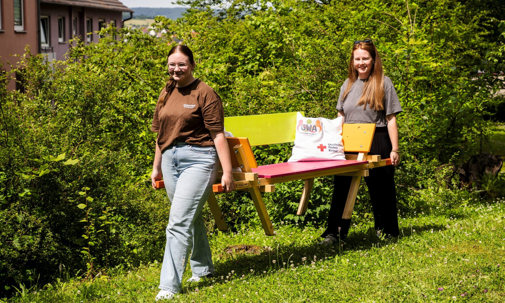 Zwei Frauen tragen eine bunt bemalte Holzbank über eine Rasenfläche. Im Hintergrund Büsche und ein Gebäude.