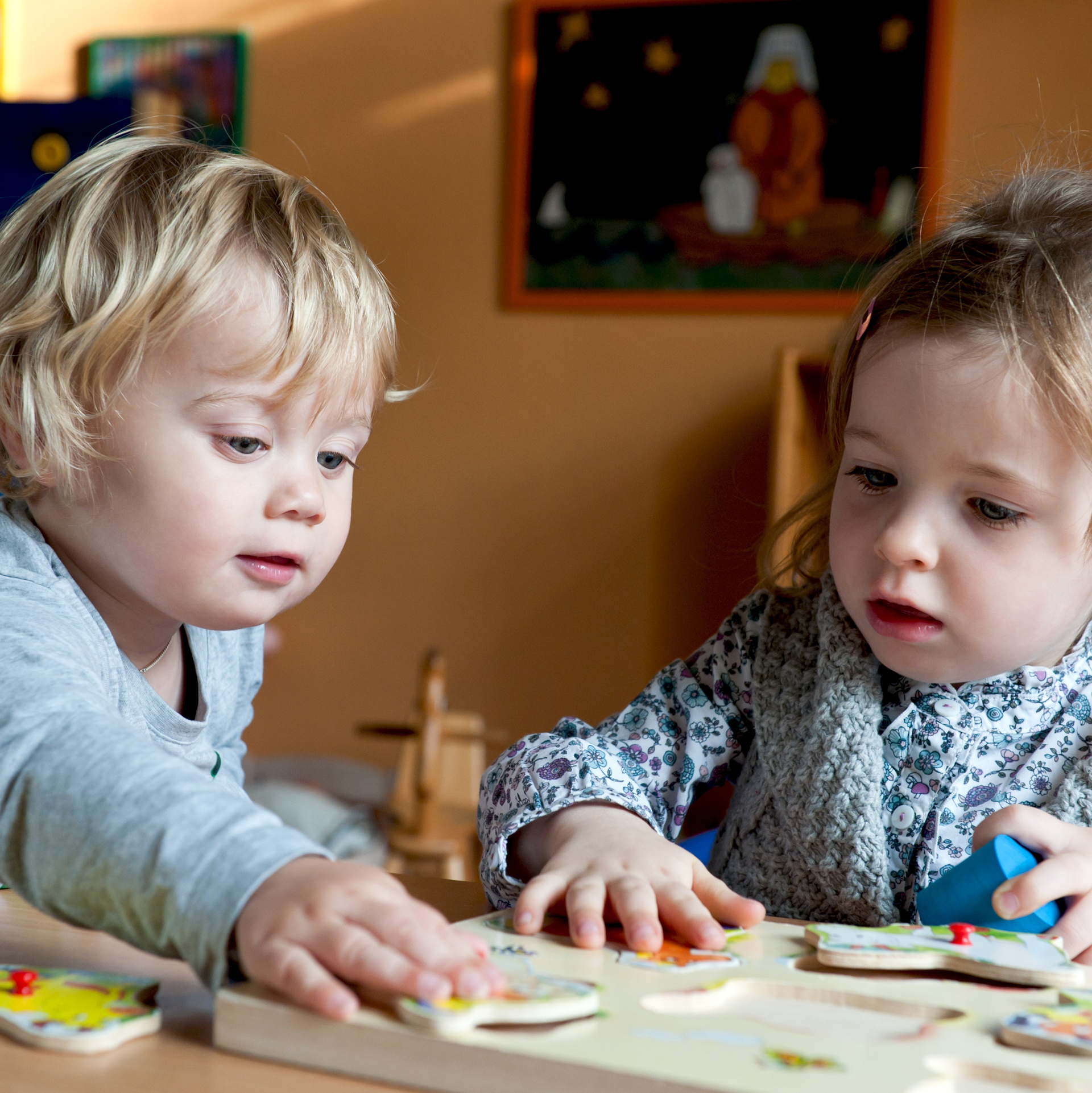 Das Bild zeigt zwei kleine Kinder - einen Jungen und ein Mädchen, die gemeinsam ein Kinderpuzzle aus Holz spielen