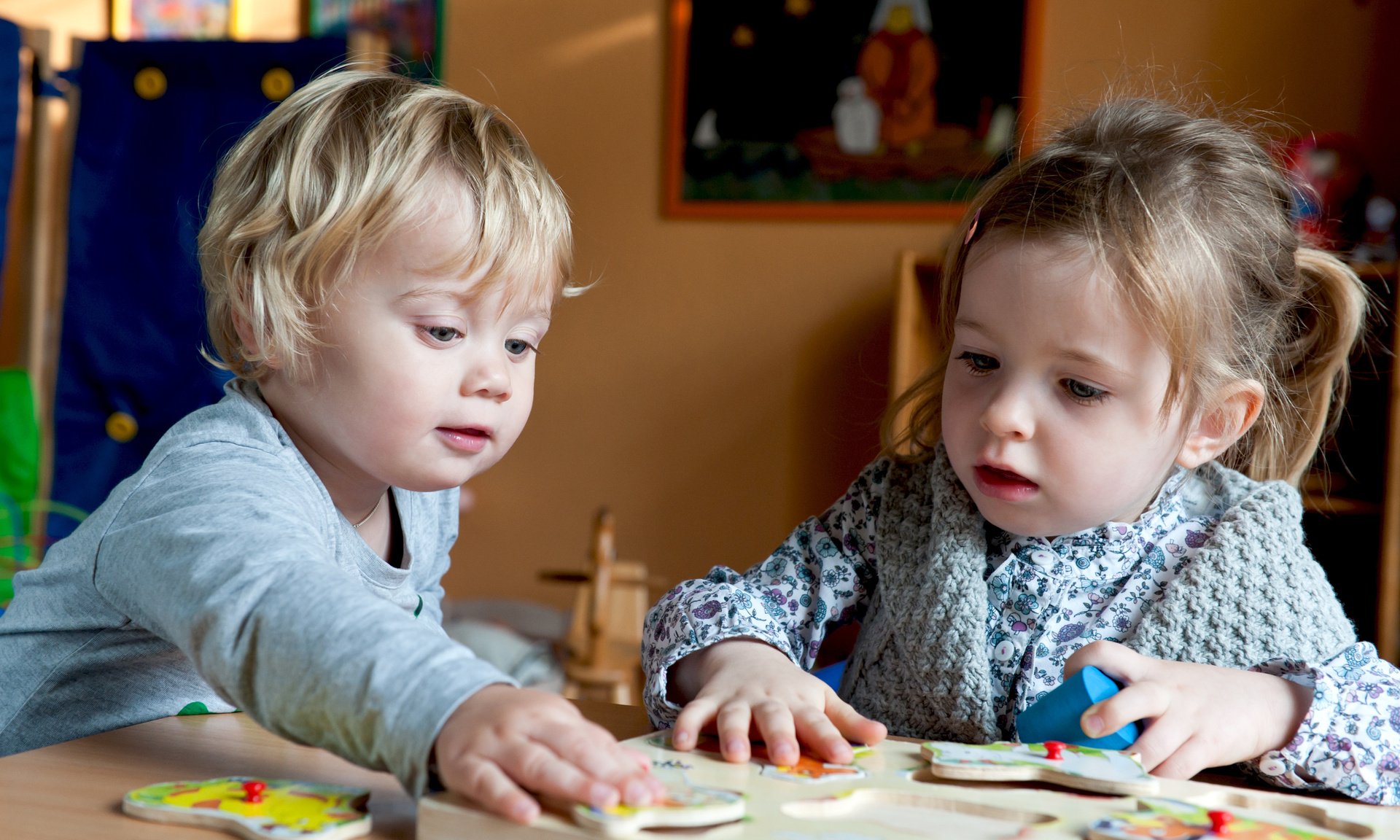 Das Bild zeigt zwei kleine Kinder - einen Jungen und ein Mädchen, die gemeinsam ein Kinderpuzzle aus Holz spielen