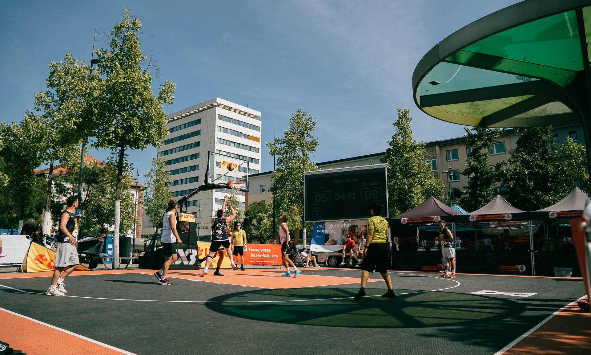 Streetbasketballcourt mit meheren Spielern in Aktion im Hintergrund Wohngebäude  