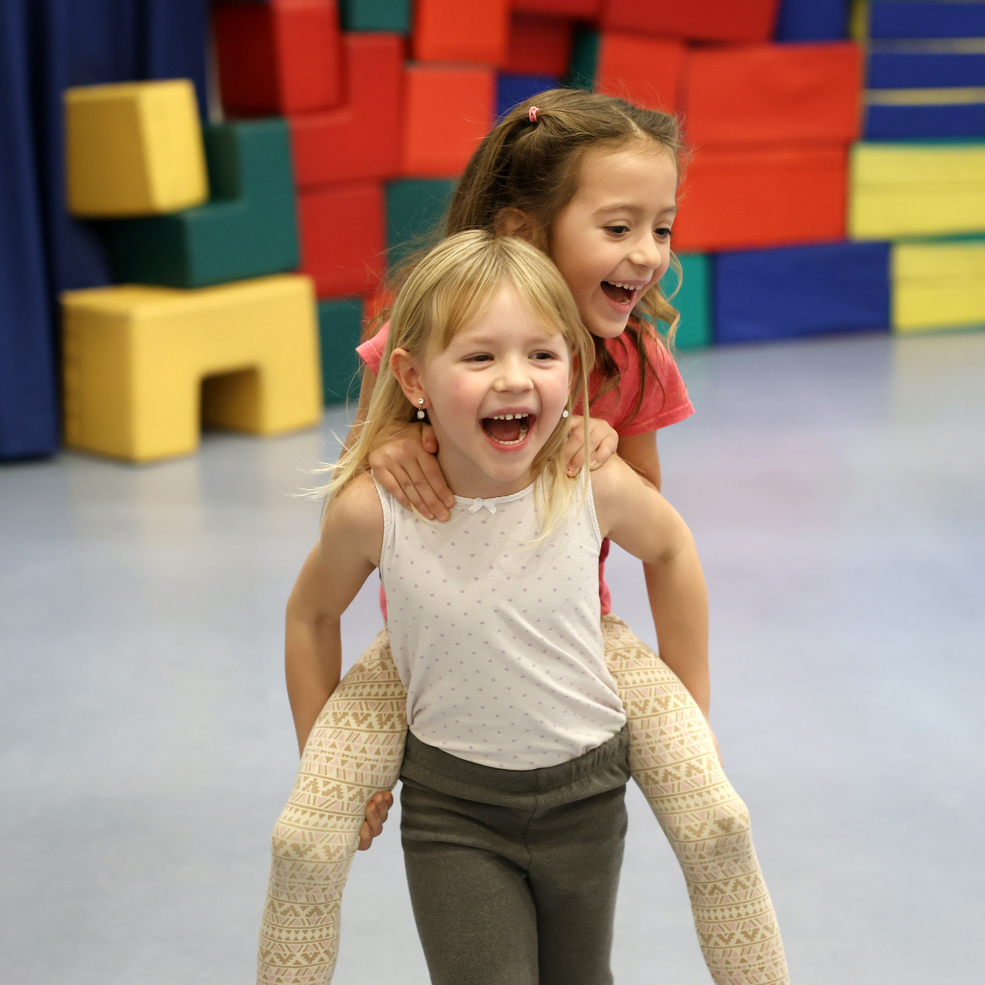 Zwei Kinder laufen in einer Sporthalle. Im Hintergrund sind Matten zu sehen. Ein Kind trägt das andere Huckepack. Kinder lachen.