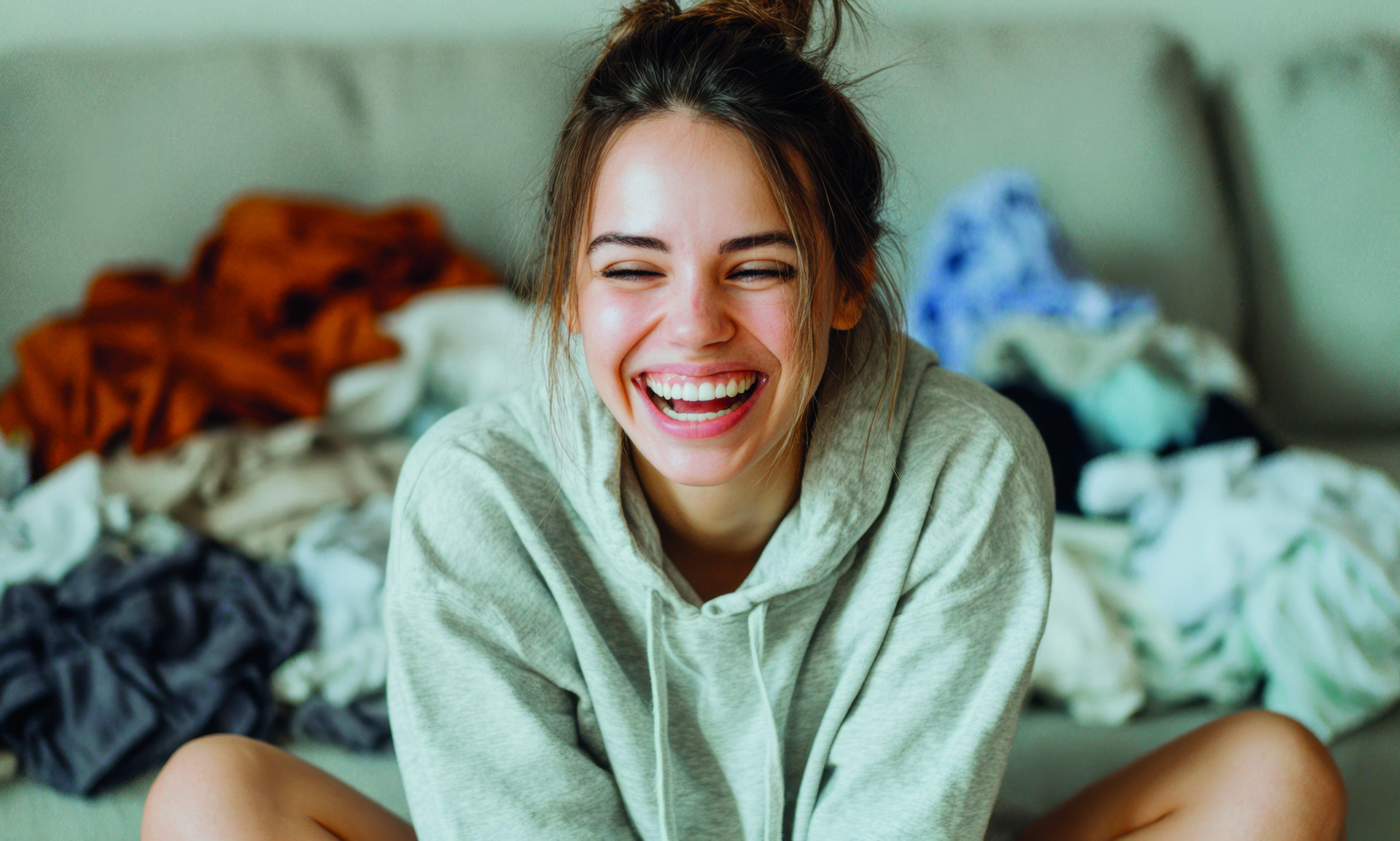 Eine lachende Frau mit braunen Haaren sitzt lachend im Schneidersitz auf einem Bett, im Hintergrund sieht man Klamotten-Stapel