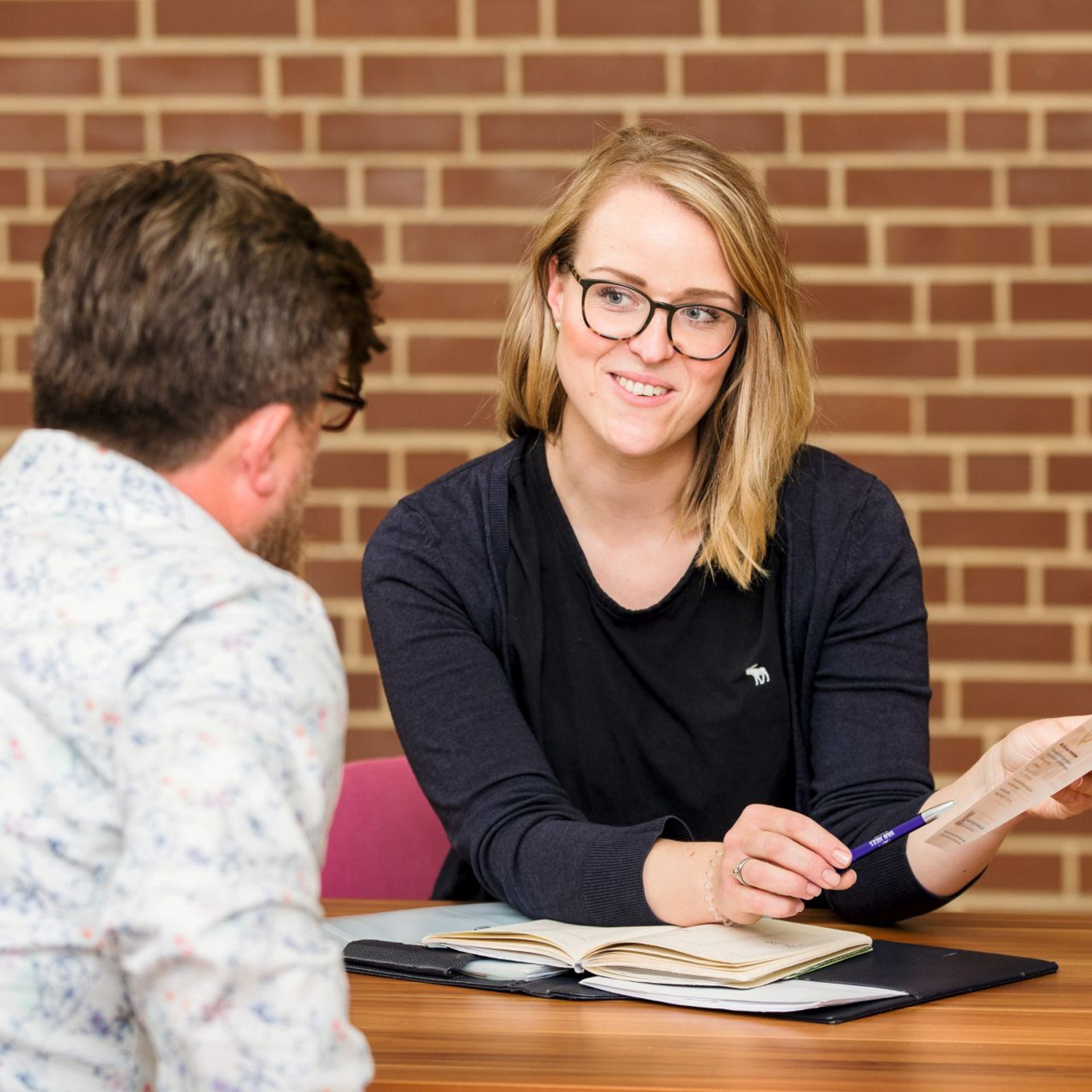 Frau sitzt vor einer rot gekachelten Wand und berät eine Person. Person sitzt mit dem Rücken zur Kamera.