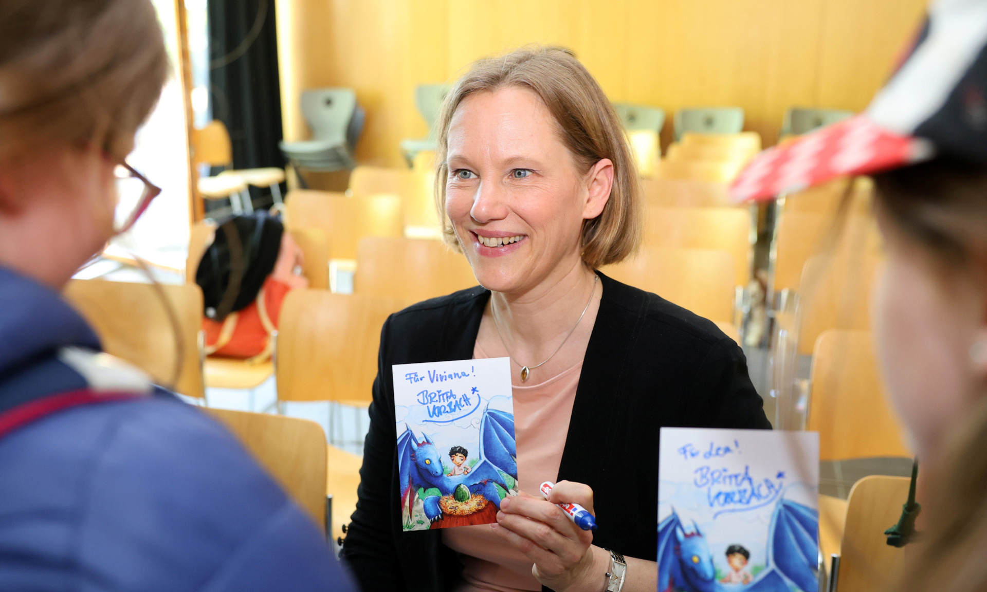 Buchautorin hält ihr Buch in der Hand und schaut seitlich in die Kamera. Im Vordergrund verschwommen zwei weitere Personen, die mit der Autorin reden