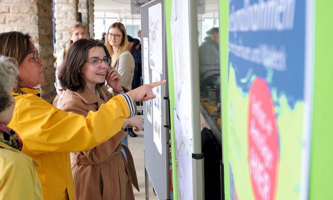 Zwei Frauen stehen vor einem Plakat und zeigen mit den Fingern darauf