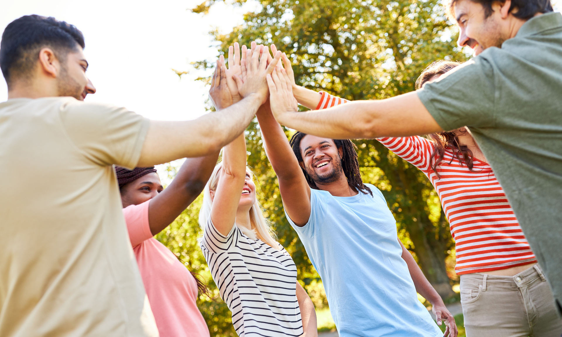 Eine Gruppe sechs junger Menschen steht draußen im Grünen und macht ein gemeinsames High-Five.
