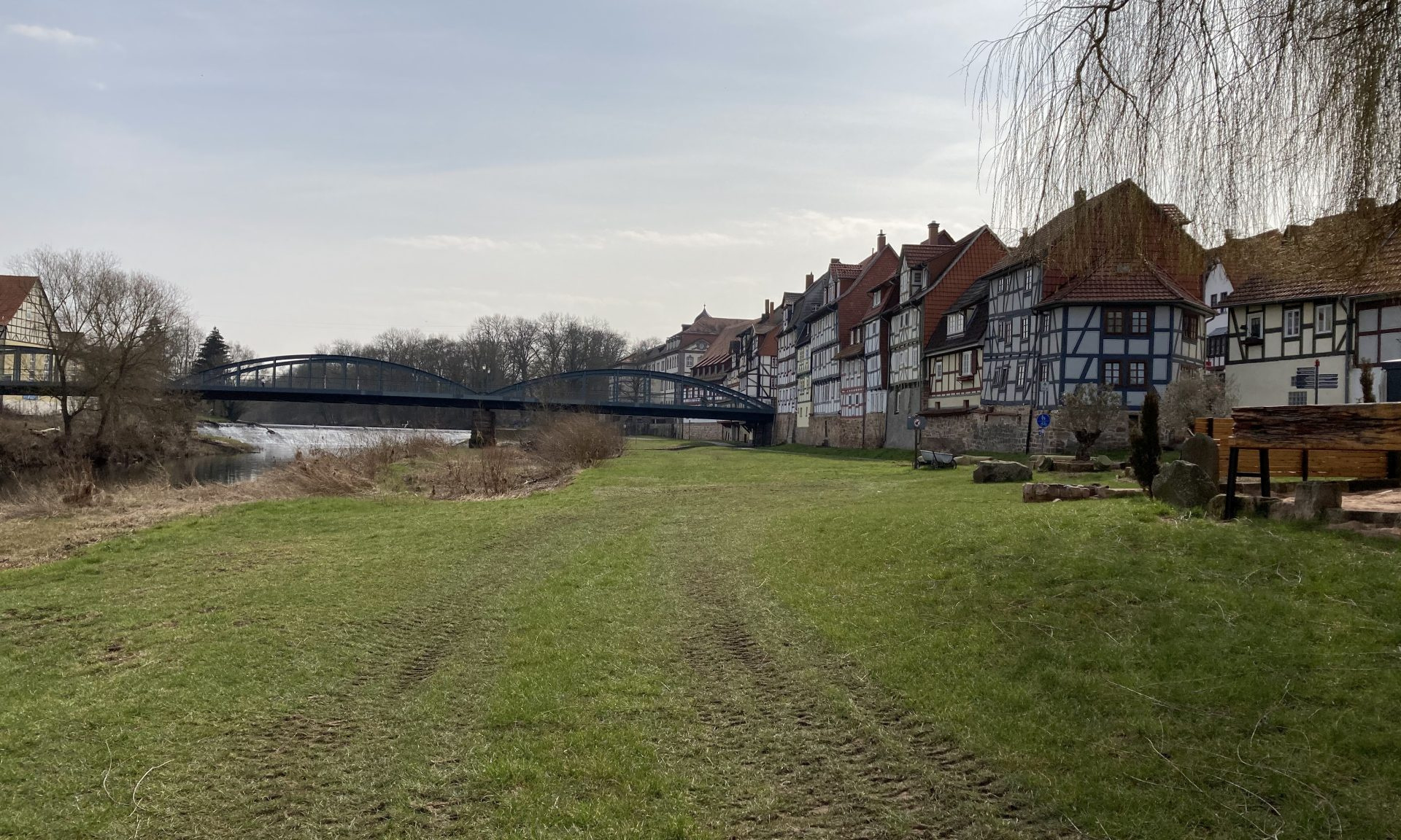 Blick auf eine ungepflegte Wiese, rechts Fachwerkhäuser, im Hintergrund eine Brücke