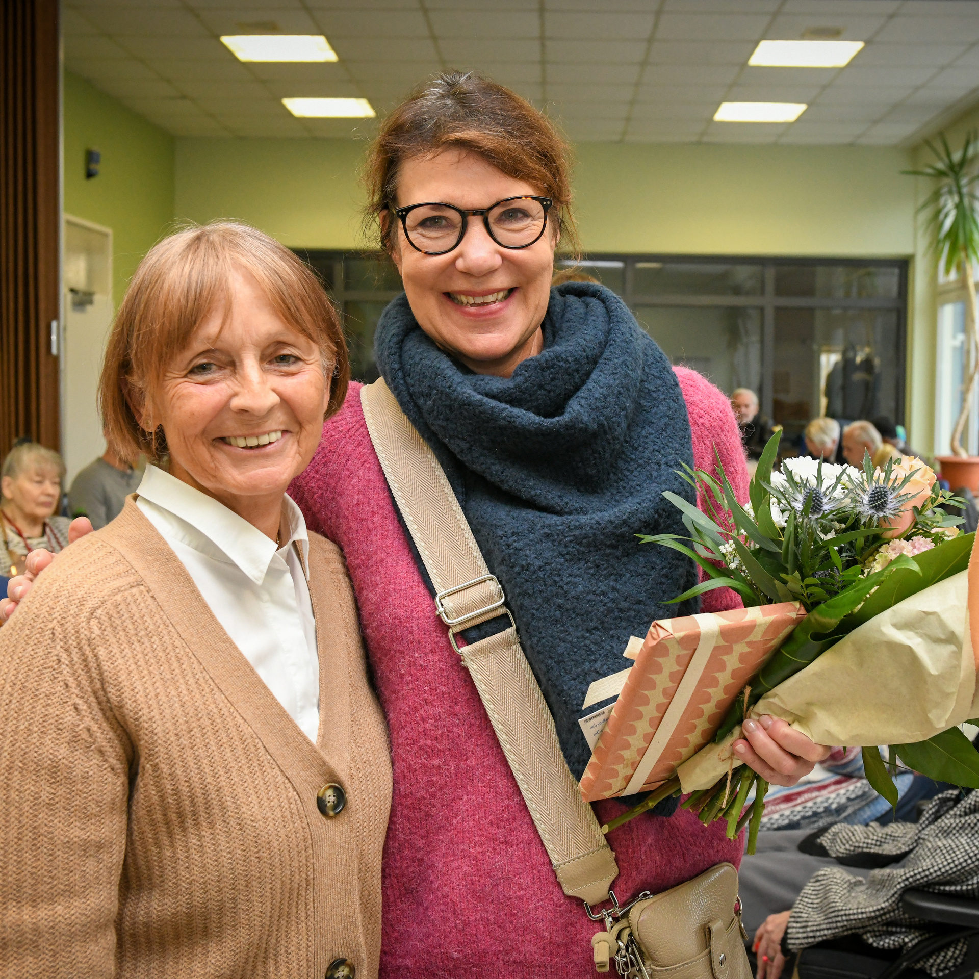 Mitarbeiterin von NHW und Leiterin vom Volksbildungswerkt steht mit einem Blumenstrauß daneben. Beide lachen in die Kamera.