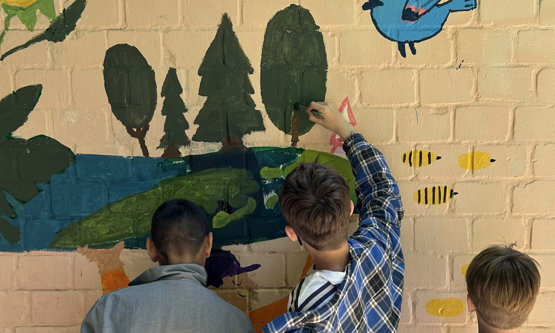 Drei Jungen von hinten zu sehen bemalen eine Wand mit Bäumen, Vögeln und anderen Tieren in grün, blau und gelb