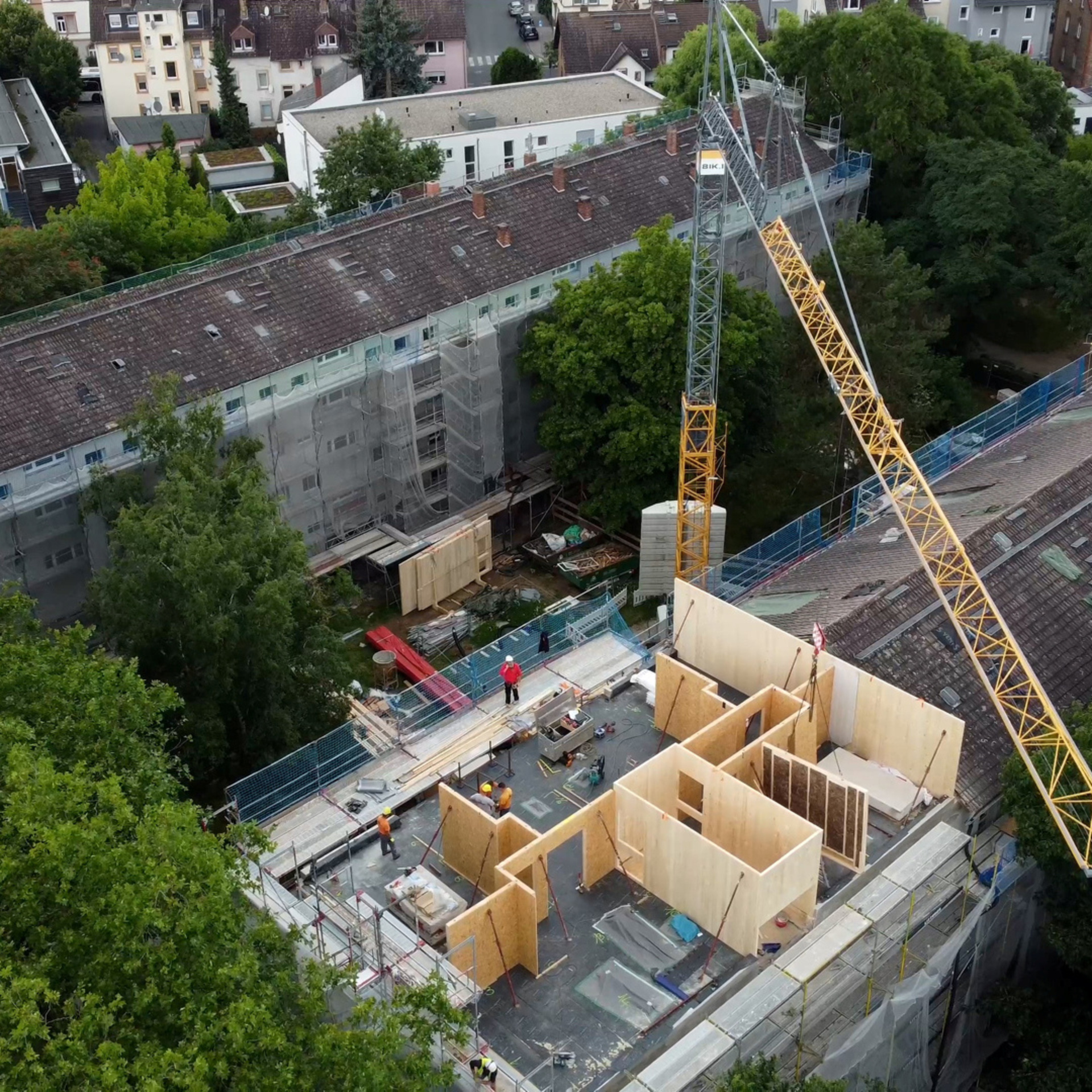 Luftaufnahme von einem Haus. Das Dach fehlt, darauf zu sehen sind Holzmodule und ein Kran.