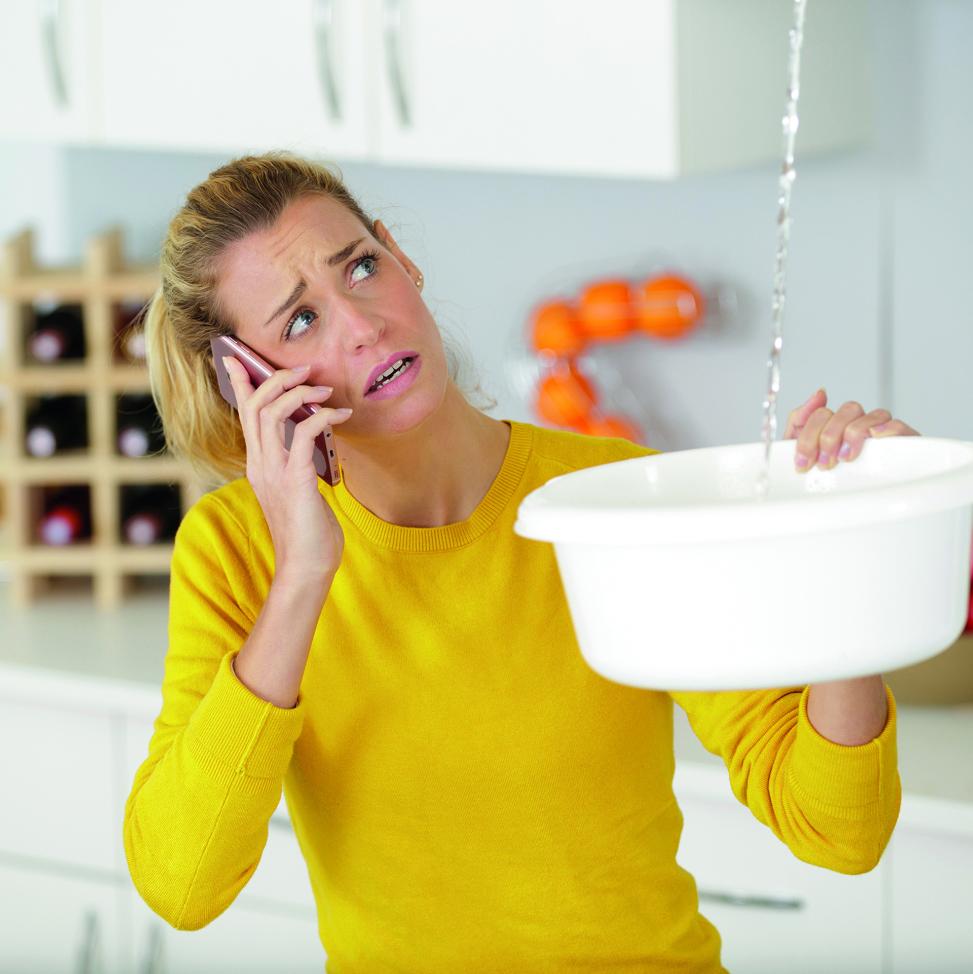 Frau hält eine Schüssel in der Hand und fängt tropfendes Wasser von der Decke auf. Hat das Handy am Ohr und ruft jemanden an