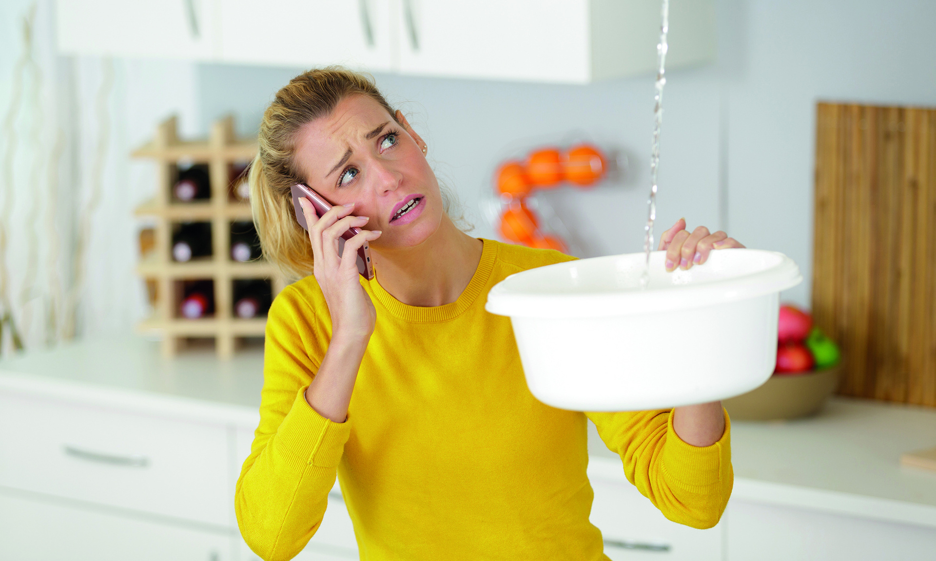 Frau hält eine Schüssel in der Hand und fängt tropfendes Wasser von der Decke auf. Hat das Handy am Ohr und ruft jemanden an
