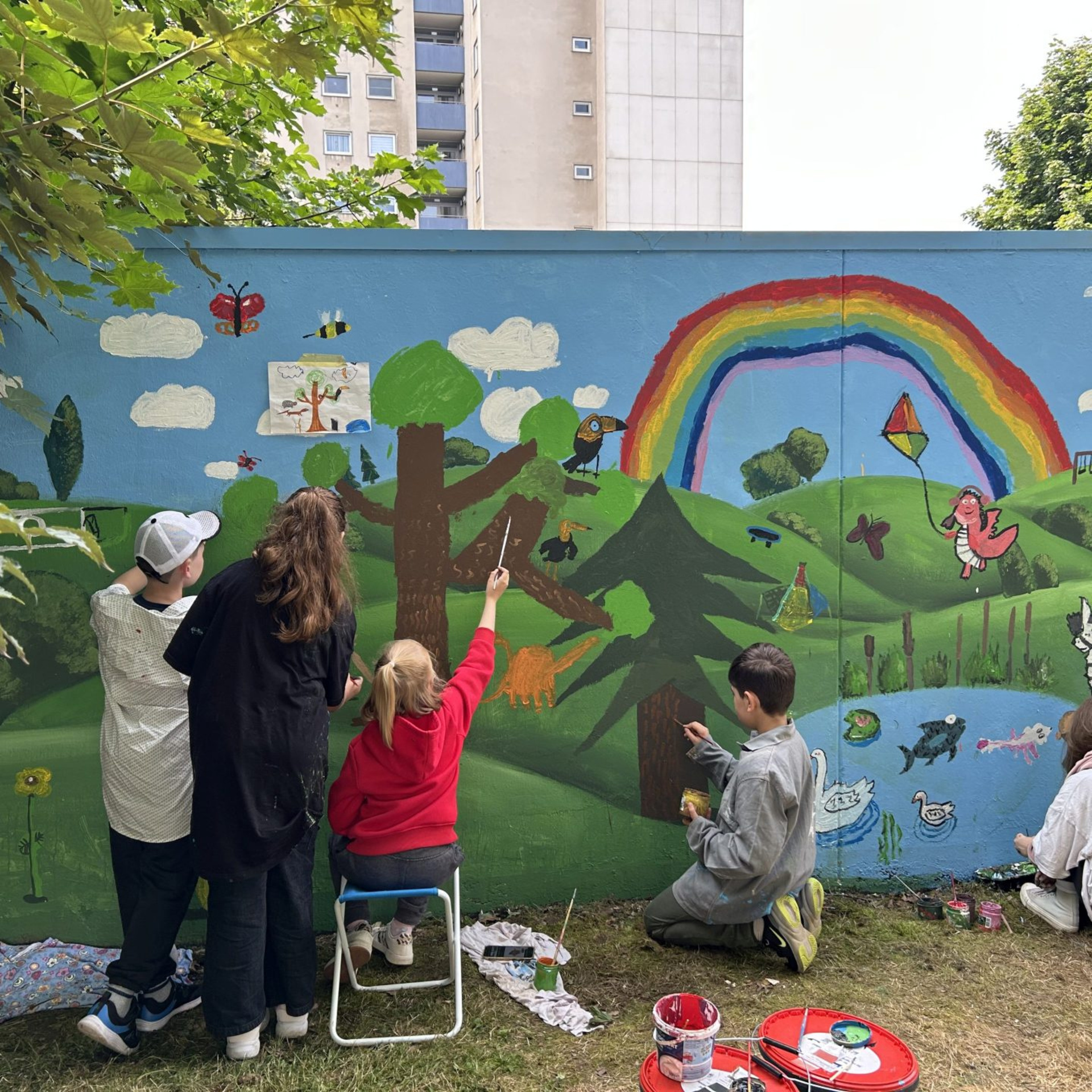 Kinder von hinten zu sehen bemalen mit bunten Farben eine Wand mit Regenbogen, Tieren, Pflanzen und einem See.