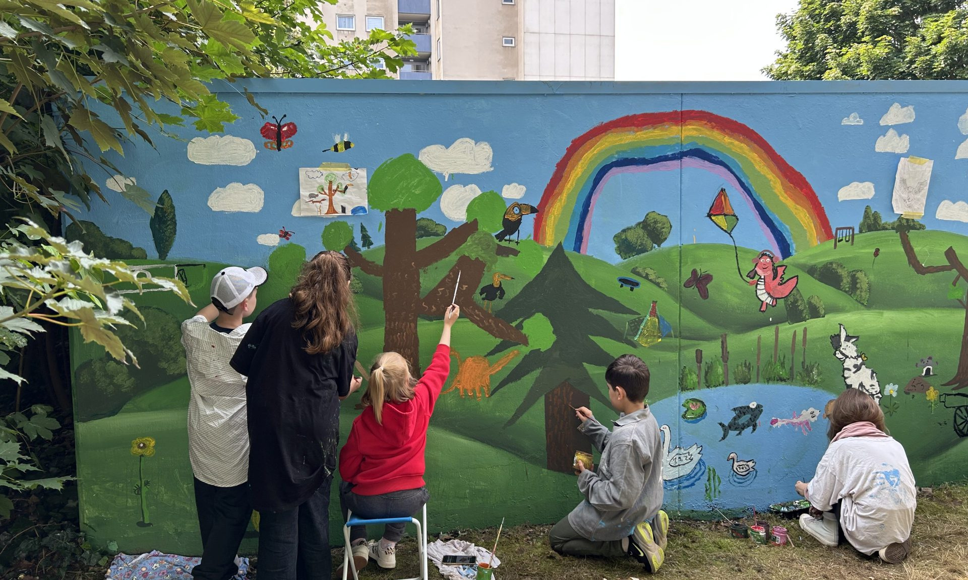 Kinder von hinten zu sehen bemalen mit bunten Farben eine Wand mit Regenbogen, Tieren, Pflanzen und einem See.