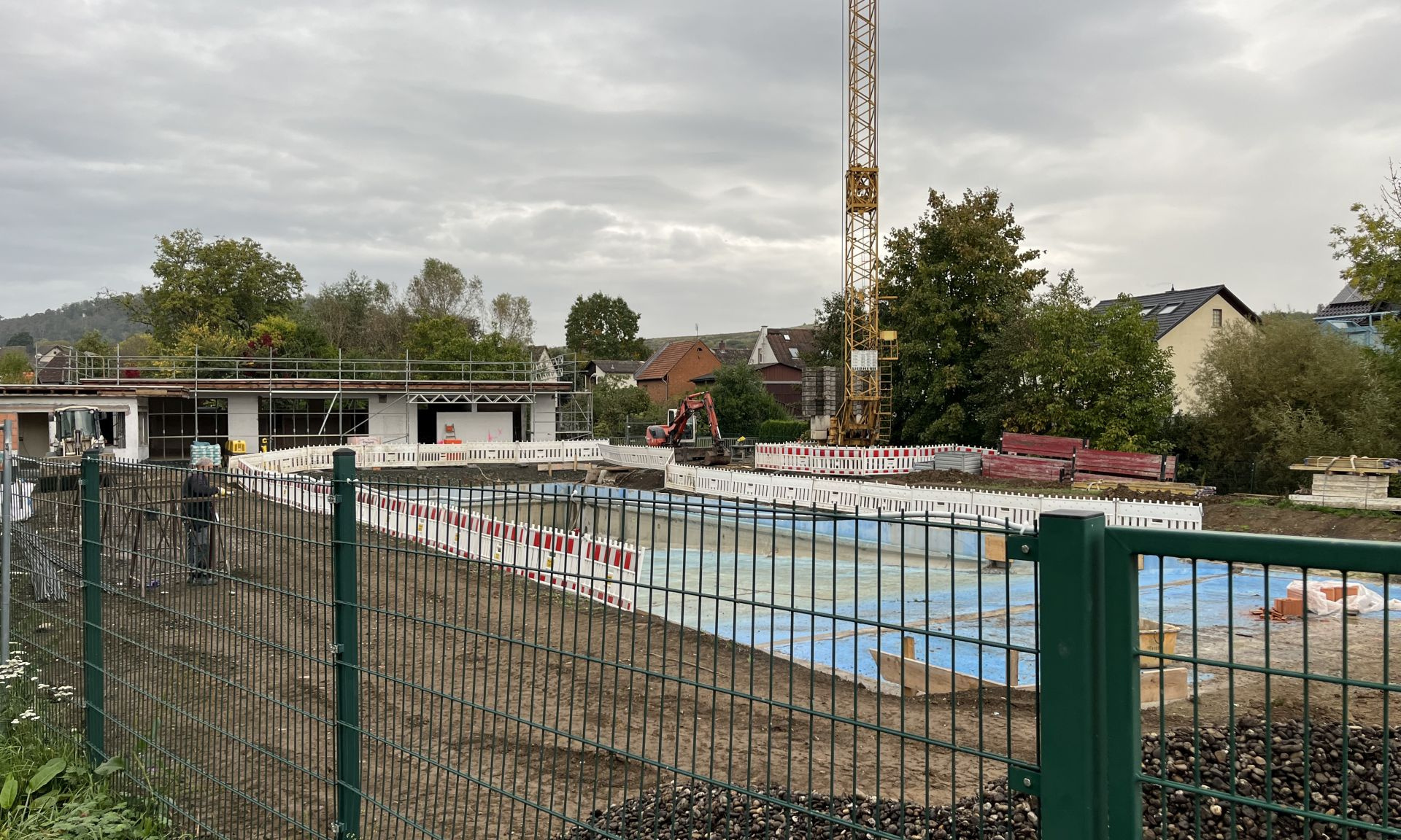 Blick auf das Freibad von Großen-Buseck mit Gerüst und Baustellenabsperrungen, im Vordergrund ein grüner Zaun, im Hintergrund ein Kran