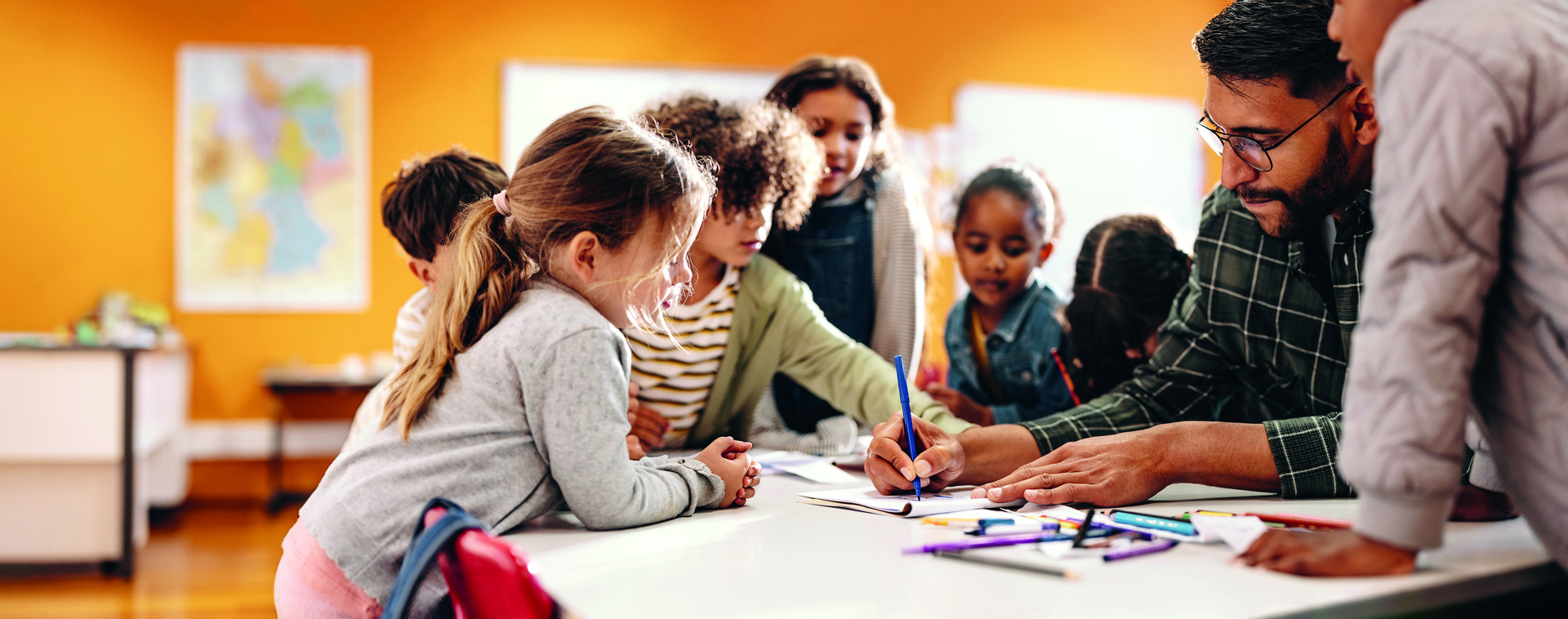 Kinder sind um einen Tisch rum und schauen in ein Buch. Erwachsene Person hält Buch auf.