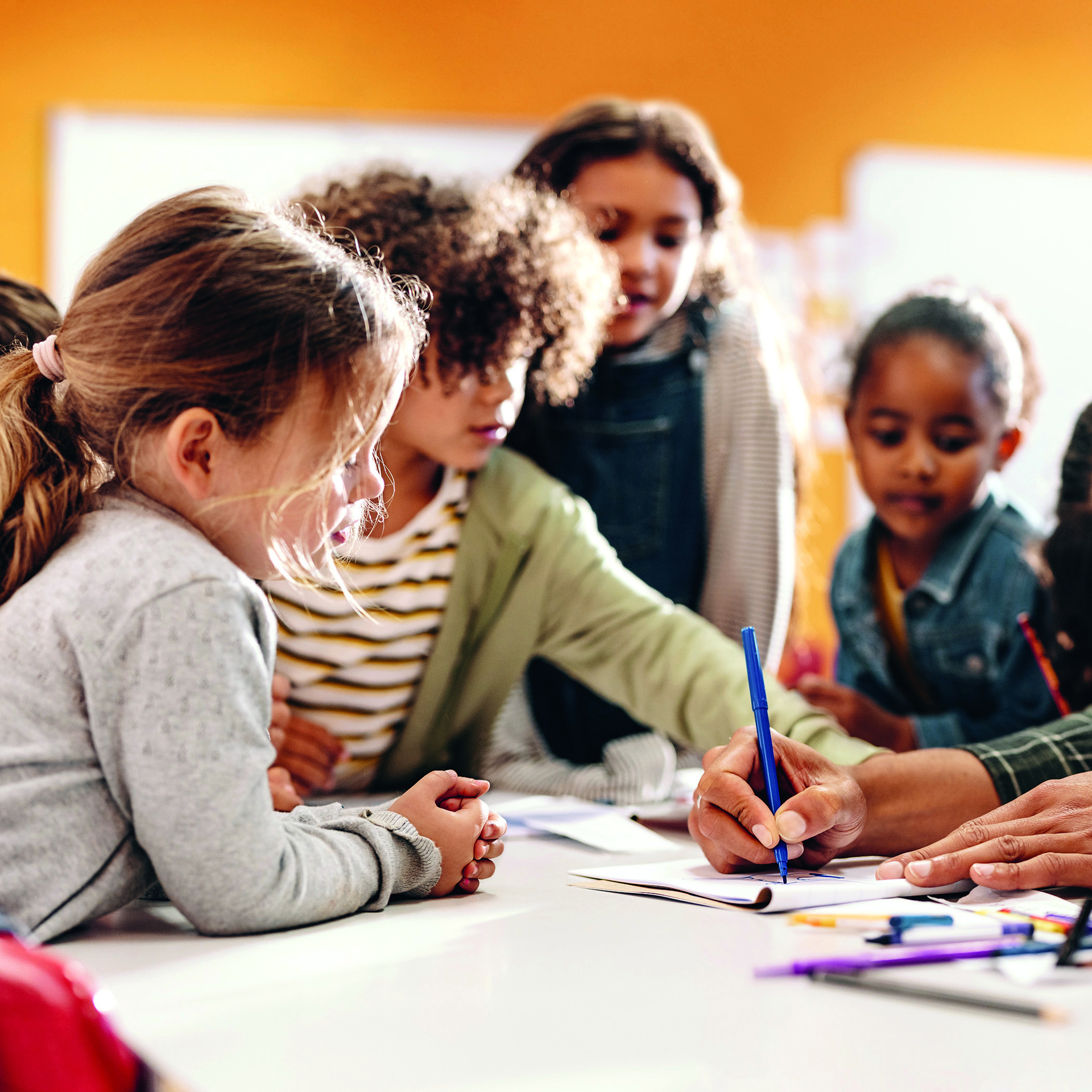 Kinder sind um einen Tisch rum und schauen in ein Buch. Erwachsene Person hält Buch auf.