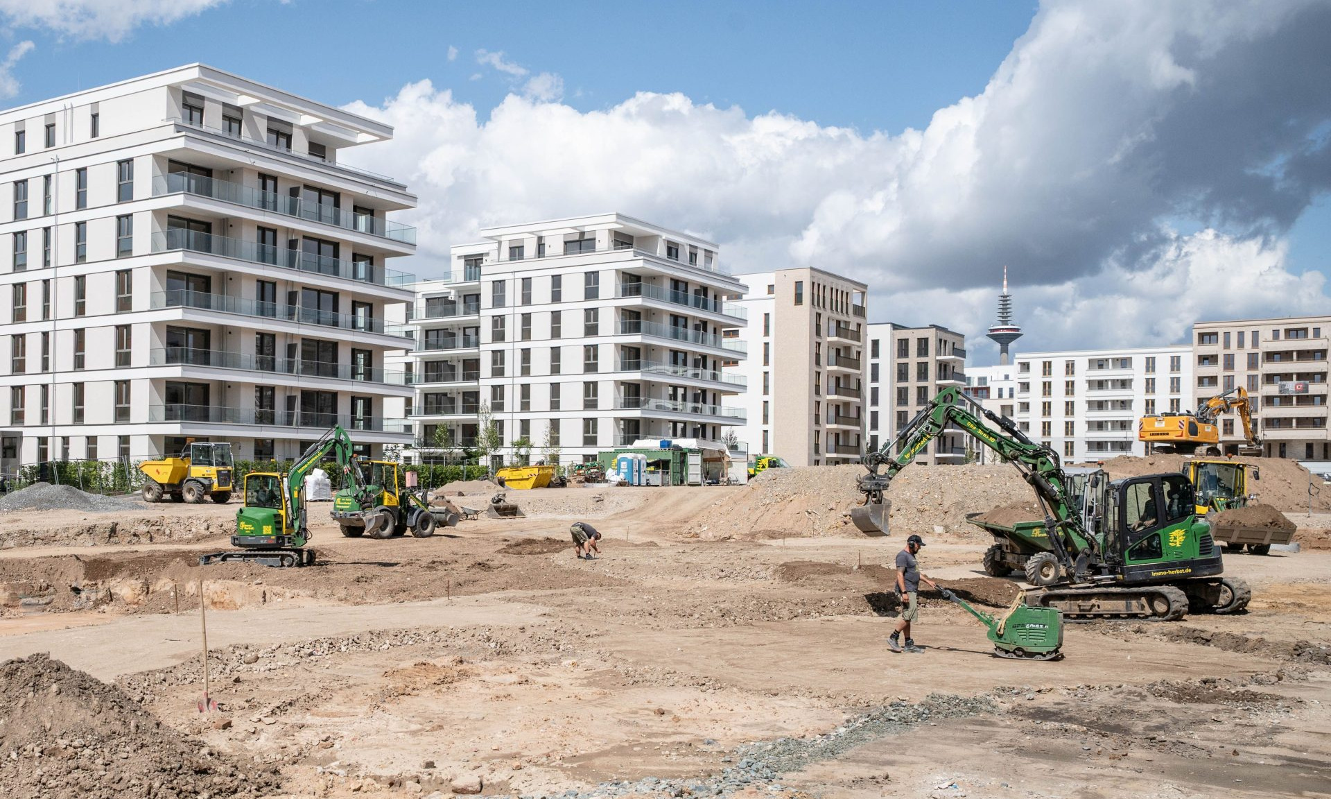 Eine Baustelle mit weitgehend fertiggestellten Neubauten im Hintergrund, im Vordergrund eine riesige Baugrube, Baustellenfahrzeuge und Bauarbeiter.