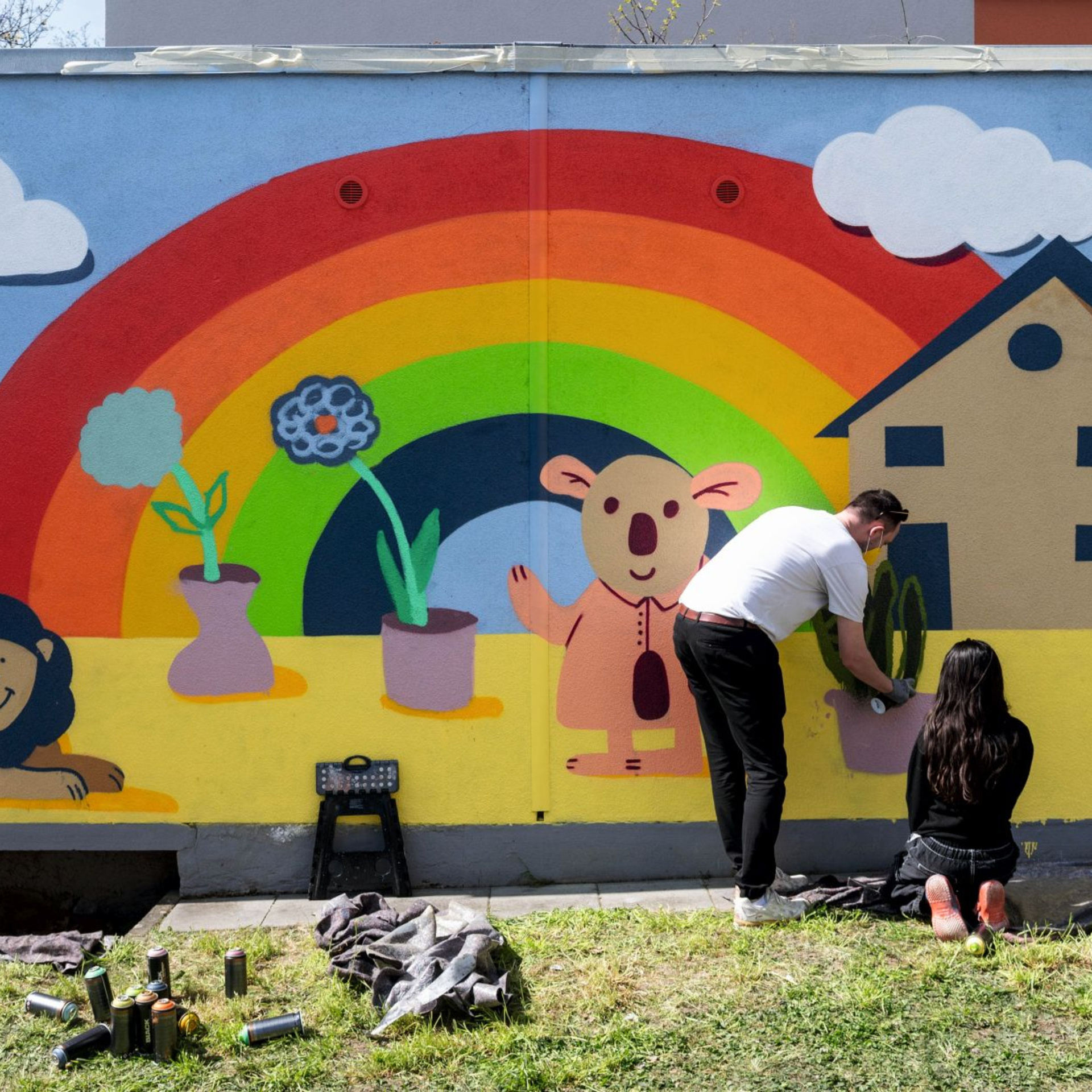 Wand mit einem Graffiti. Regenbogen mit Haus und Blumen im Topf, ein Löwe. Mädchen sprüht und ein Anleiter steht dabei.
