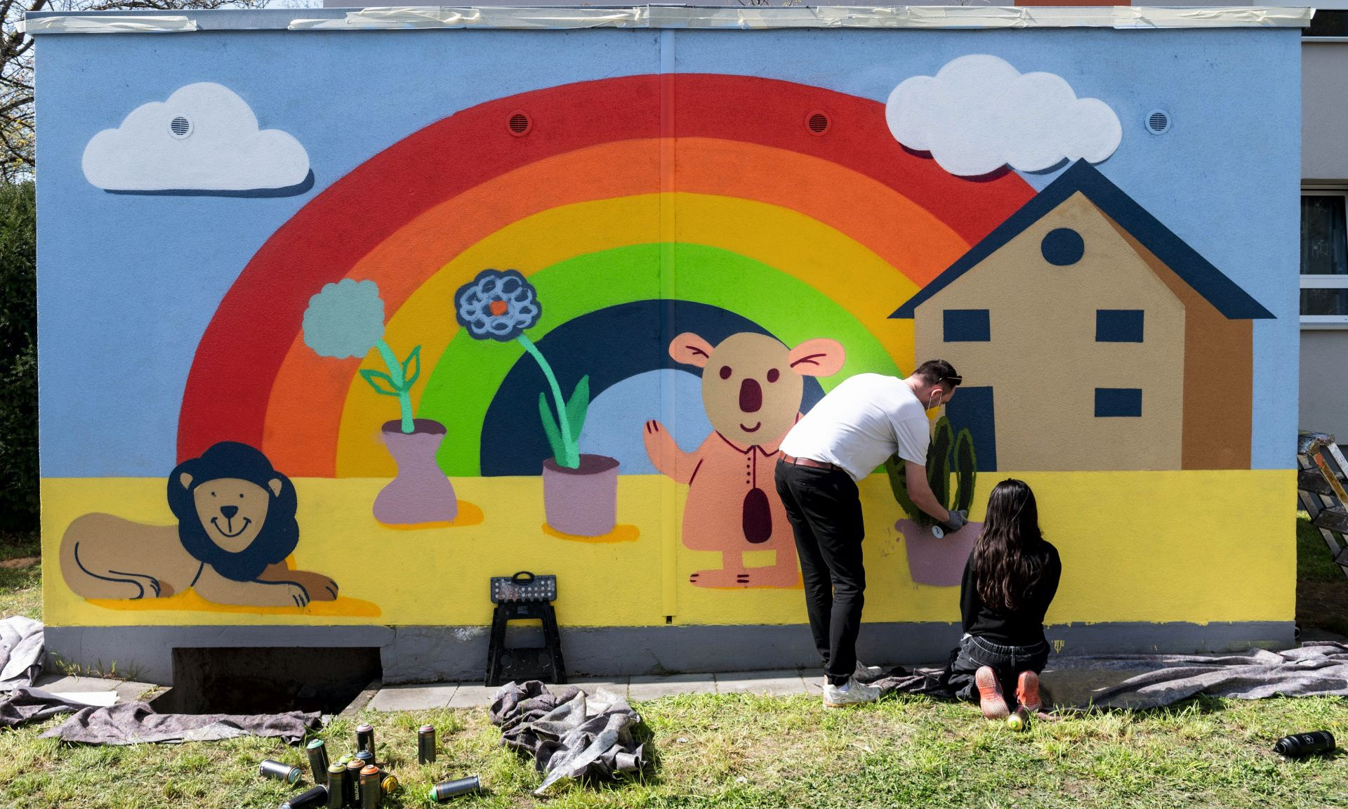 Wand mit einem Graffiti. Regenbogen mit Haus und Blumen im Topf, ein Löwe. Mädchen sprüht und ein Anleiter steht dabei.