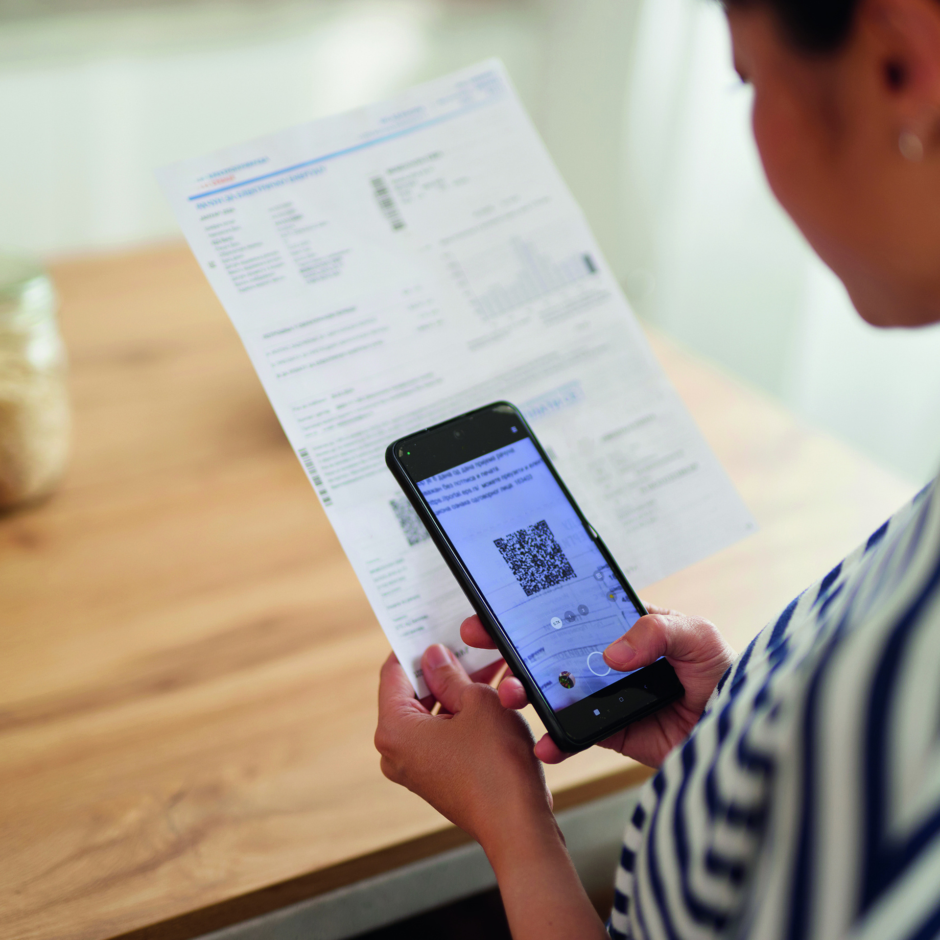 Eine Frau hält ein Handy in der Hand und scannt damit einen QR-Code, der sich auf einem DIN-A4-Papierschrieben befindet.