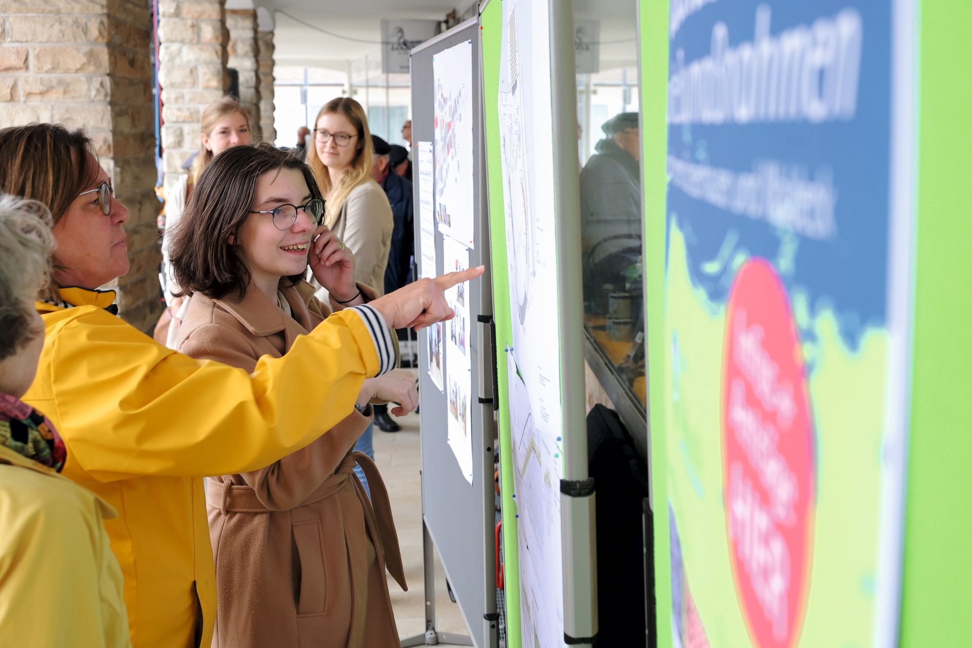Zwei Frauen stehen vor einem Plakat und zeigen mit den Fingern darauf